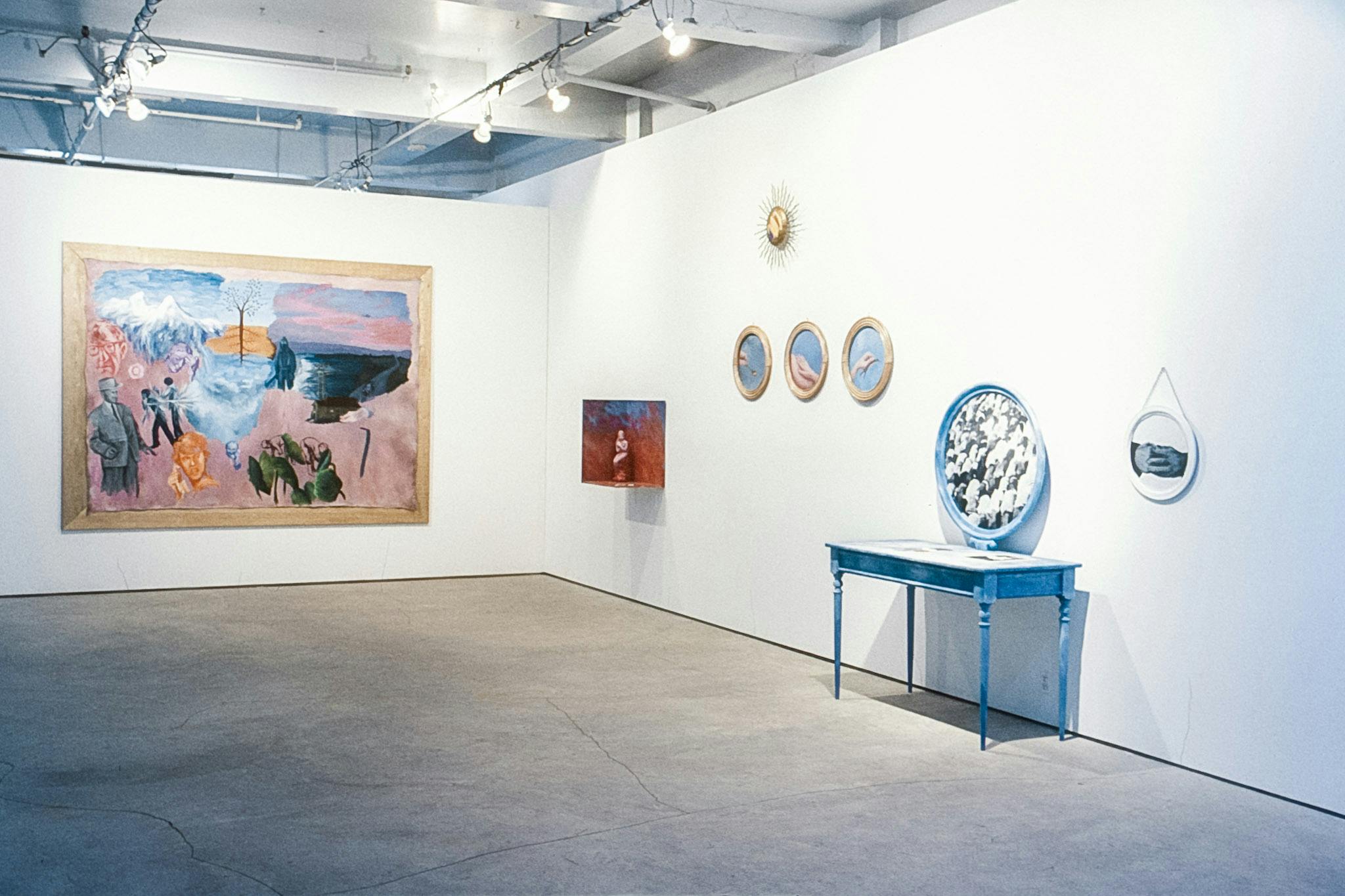 A corner in a gallery. On one wall, there is a large colourful painting in a gold frame. On the other wall there is a small red box with a sculpture on the wall, as well as other photos and a vanity.