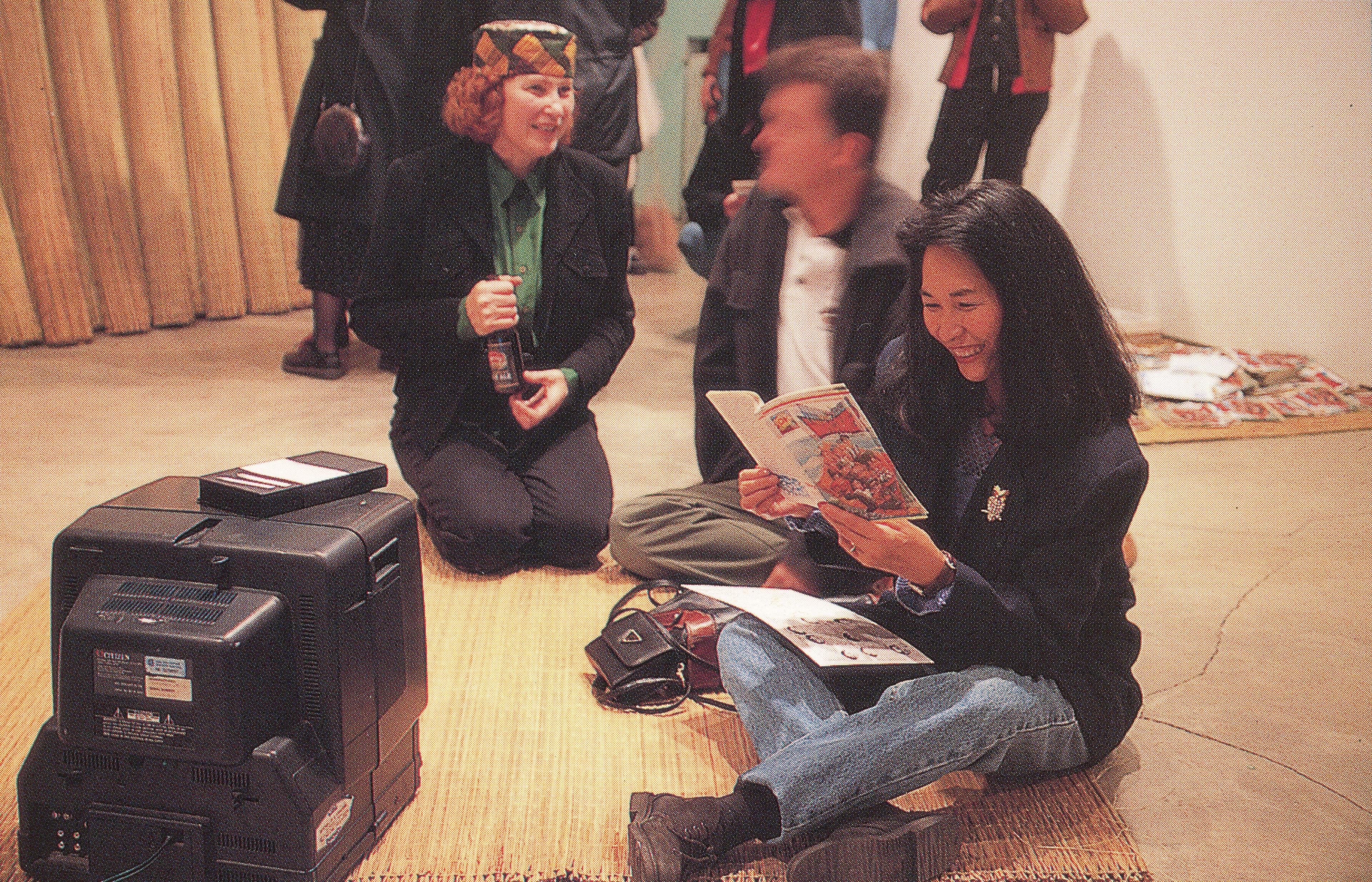 Several rush mats and a CRT TV are placed on the floor of a gallery space. A person sitting on a mat is reading a magazine made by the artist. Many more visitors are visible in the back. 