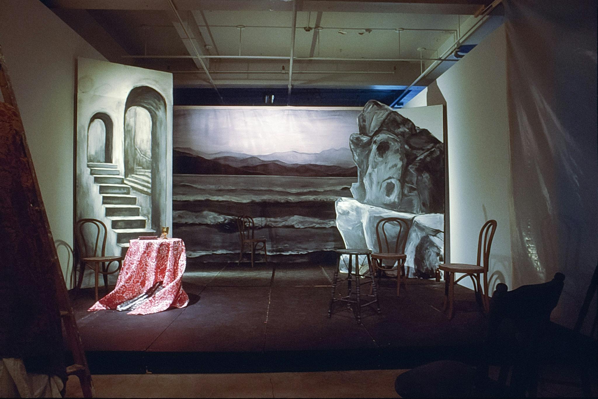 A theatre stage is set in a gallery space. A dark body of water is depicted in the stage background. Four chairs, a stool, and a table are placed on the stage distanced from each other. 