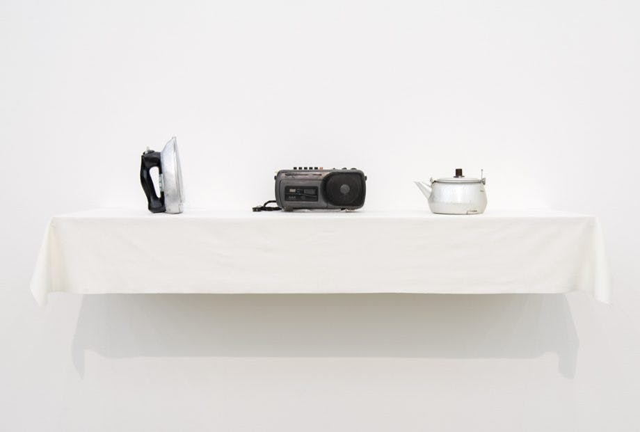 Three objects are displayed in a gallery space. From left to right, a silver iron, black portable radio, and silver kettle are placed on a shelf mounted to a white gallery wall. 
