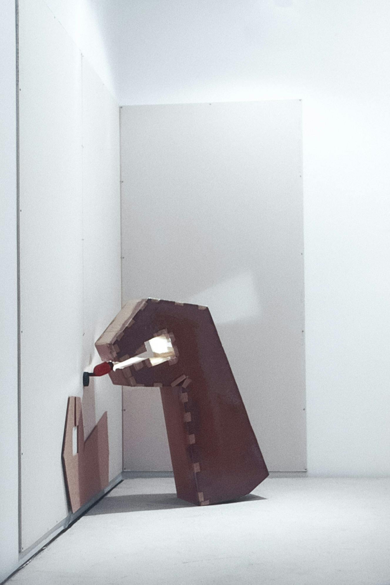 The corner of a gallery with white floors and walls. There are 3 grey panels on the walls. On 1 panel there is a flat, wrench-shaped cardboard piece. This shape is repeated in 3D, leaning on the wall. 