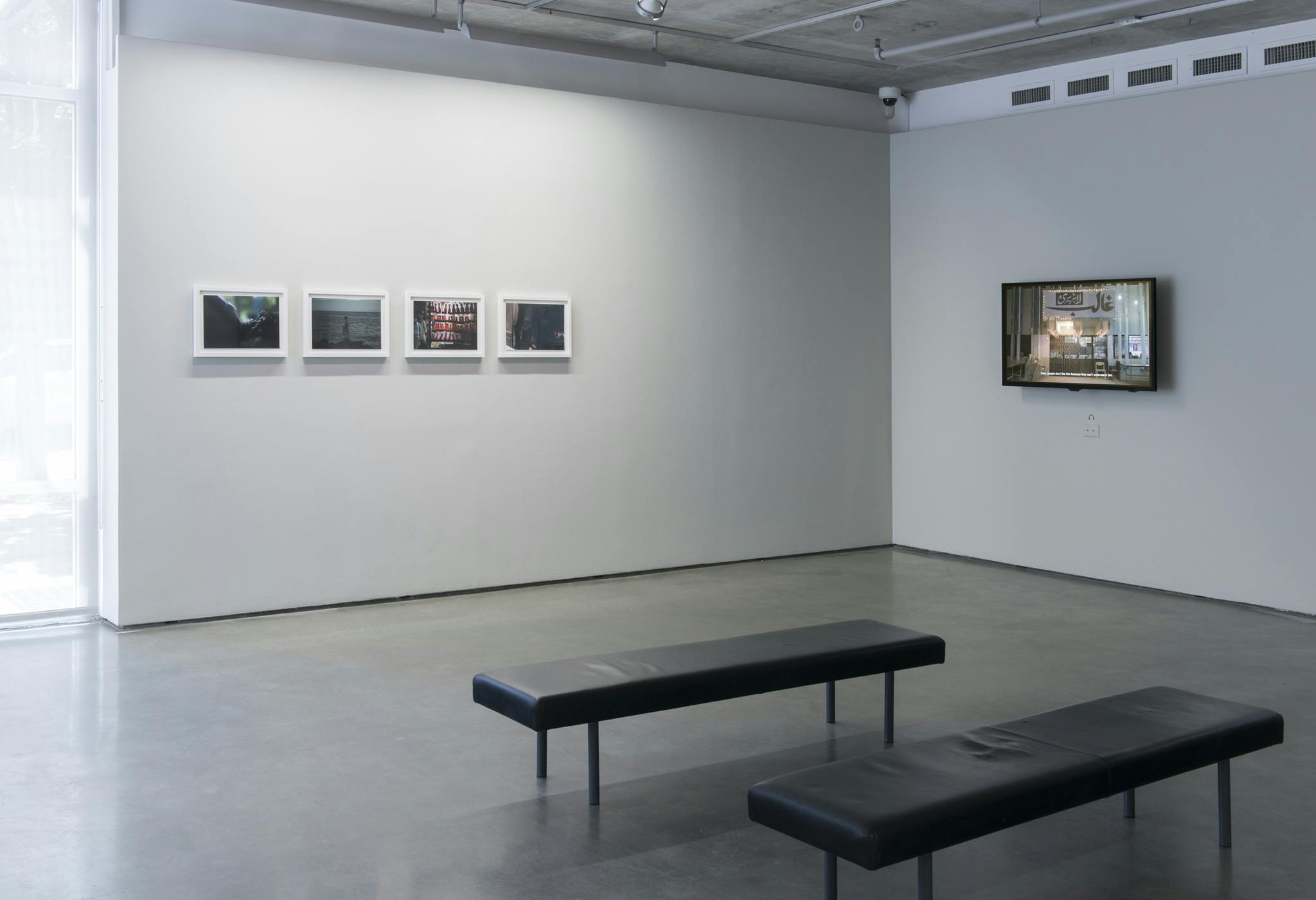 Four framed photographs and one monitor hang on two walls. On the left wall there are four framed photographs, on the right wall there is one monitor. Two benches sit in the middle of the room.