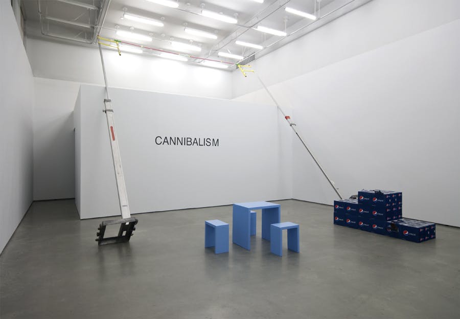 A structure resembling a pole vault leans against a wall with the word CANNIBALISM displayed in black text. On the floor, cases of Pepsi are arranged as a three tier podium near a blue table and stools.