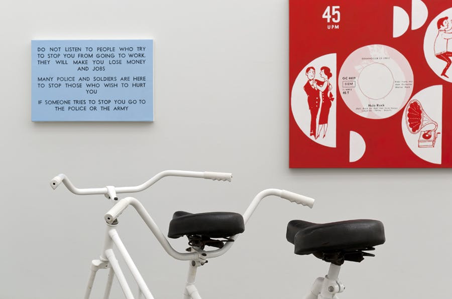 Detail image of an installation by Roy Arden. In this image, the top part of a white bike is visible below two graphic design works mounted on the wall; one is blue and the other is red.