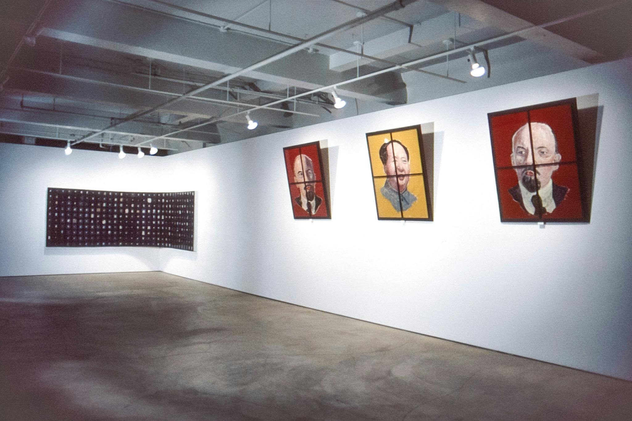A photo of an installation view in the gallery. A set of three paintings shows the head of Mao Zedong in a yellow background in between two paintings of the head of Vladimir Lenin in a red background. 
