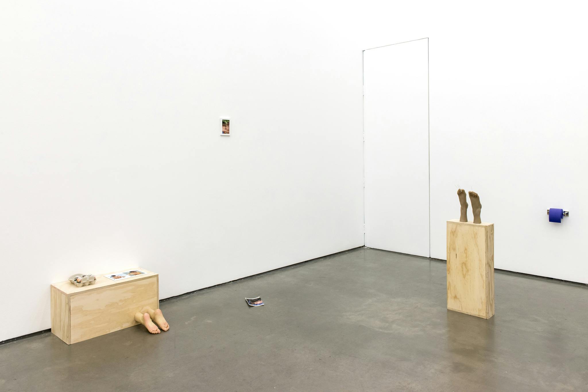 Two sculptures sit on the floor of a gallery space. The sculptures are made of wooden rectangular boxes with prosthetic feet protruding out of them. Four photographs are placed in various spots. 