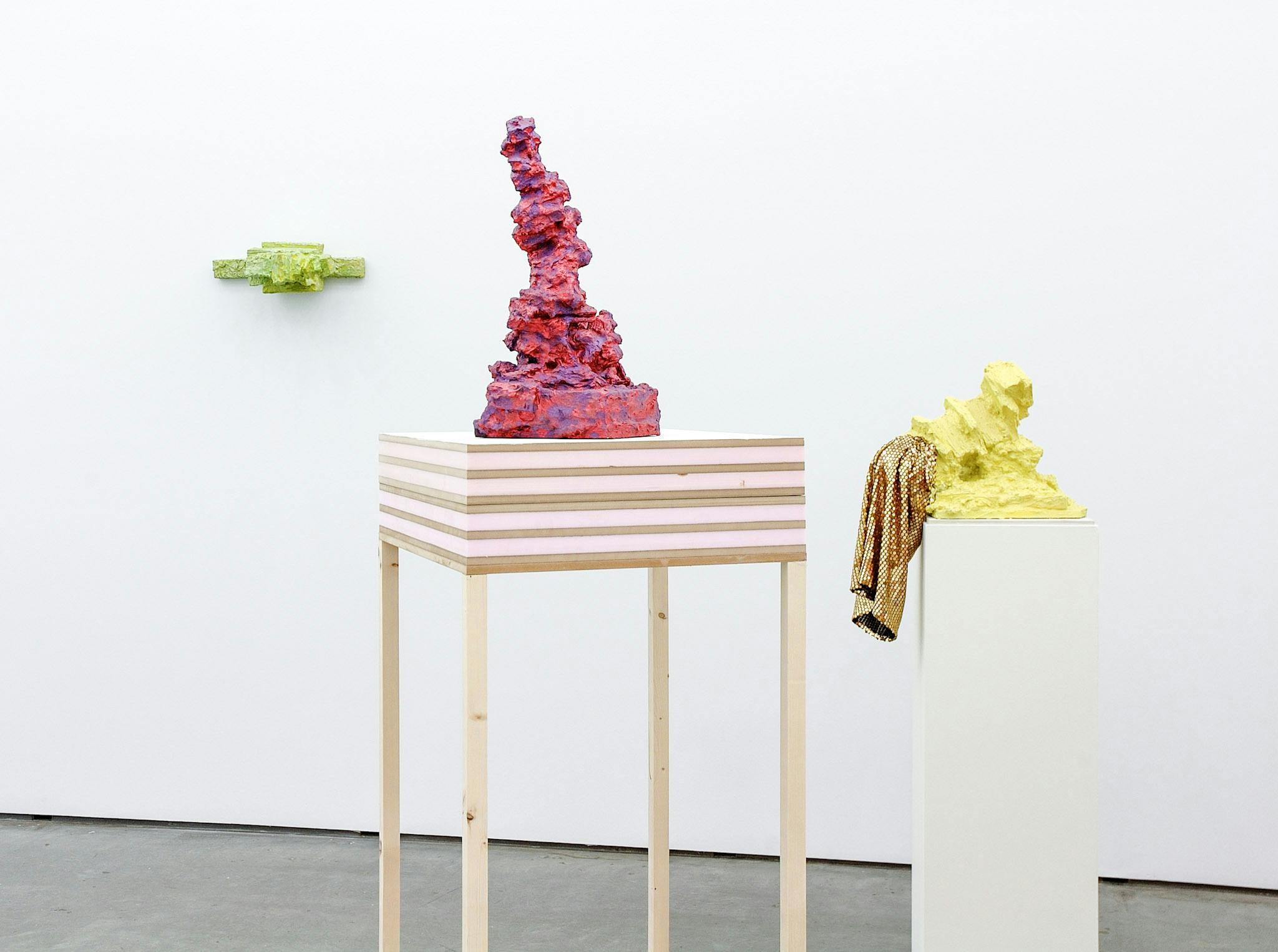 Three artworks in a gallery. One is narrow and green, mounted on the wall. One is red and purple, resting on wood and foam. The third is yellow with gold sequined fabric on top, resting on a plinth.