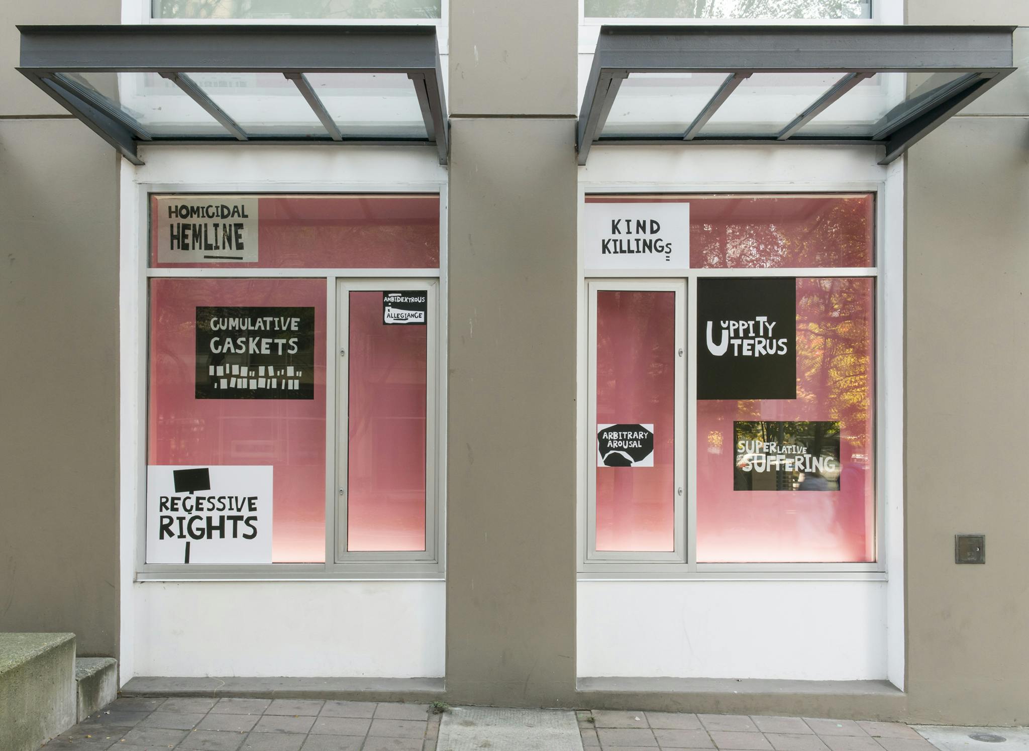 Two ground level façade windows at CAG. They each have a pink backdrop and black and white text-based vinyl prints that read “Kind Killings,” “Regressive Rights” and “Homicidal Hemiline.” 