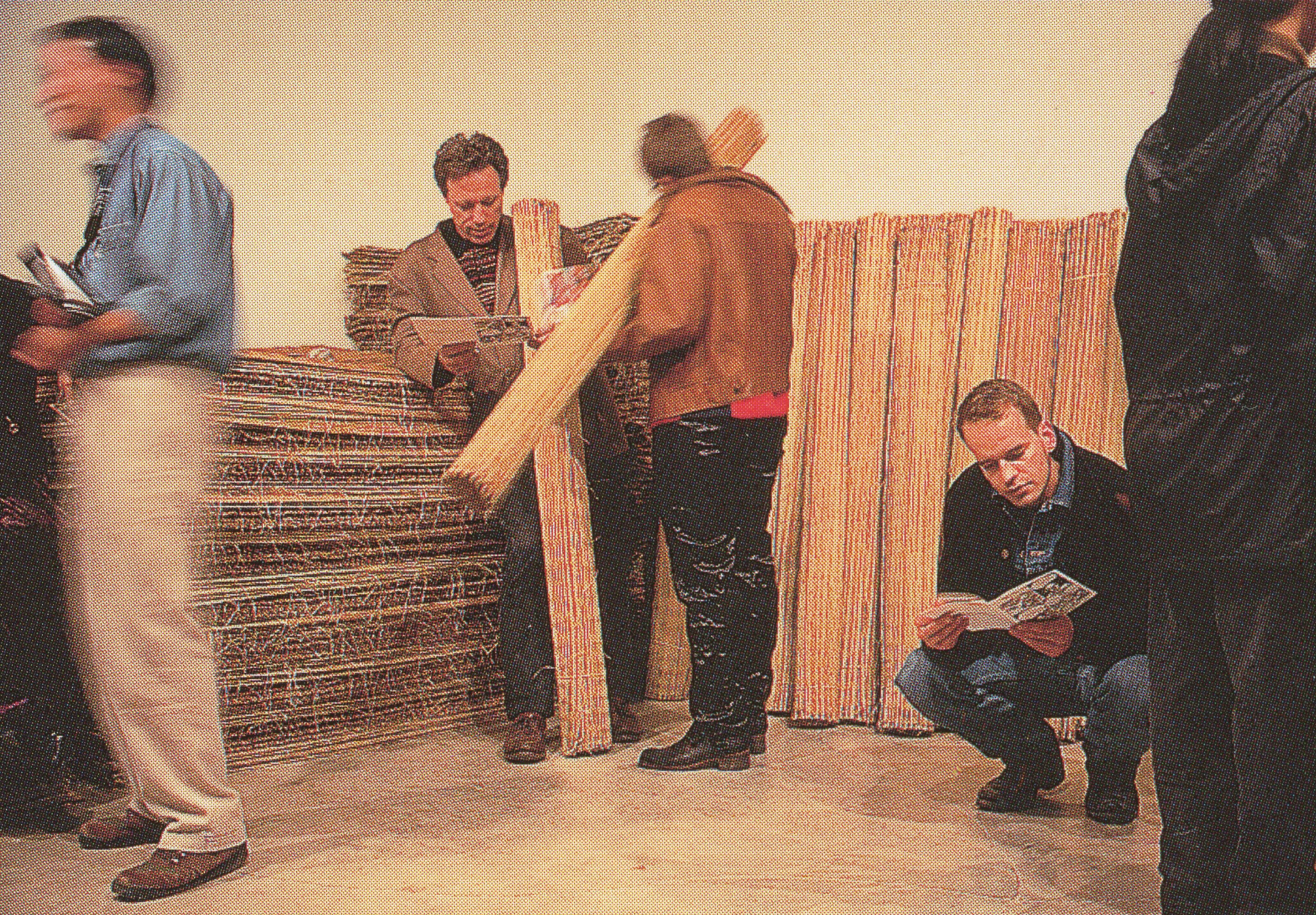 Many human-sized pillars made of straw are leaning against the gallery wall. Two standing people are reading pamphlets while holding those straw pillars. 