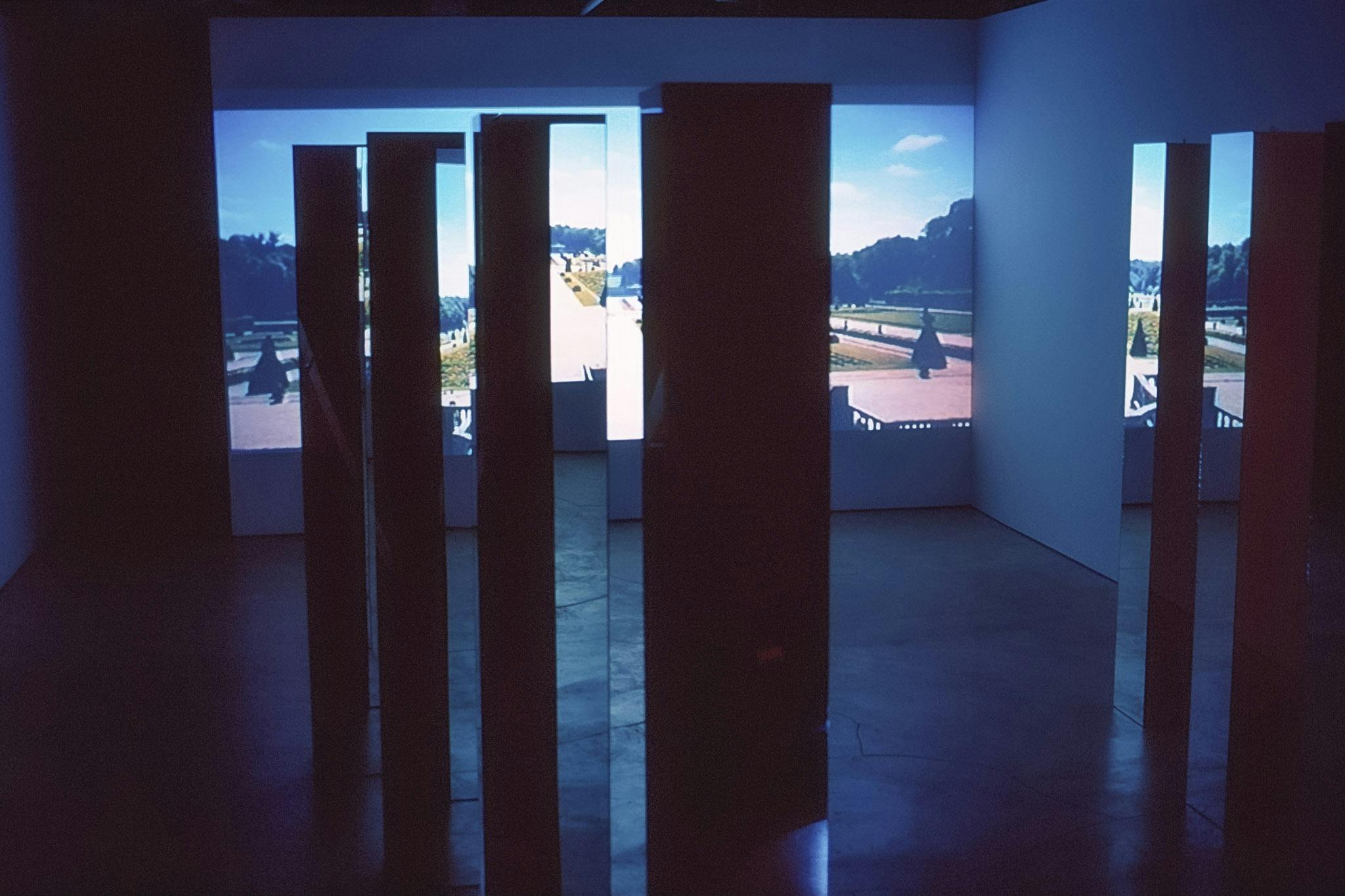 Eight large mirrored columns stand on the gallery floor, reflecting a photographed image of a garden projected on the front wall. The garden seems large and symmetrical. The gallery space is darkened.