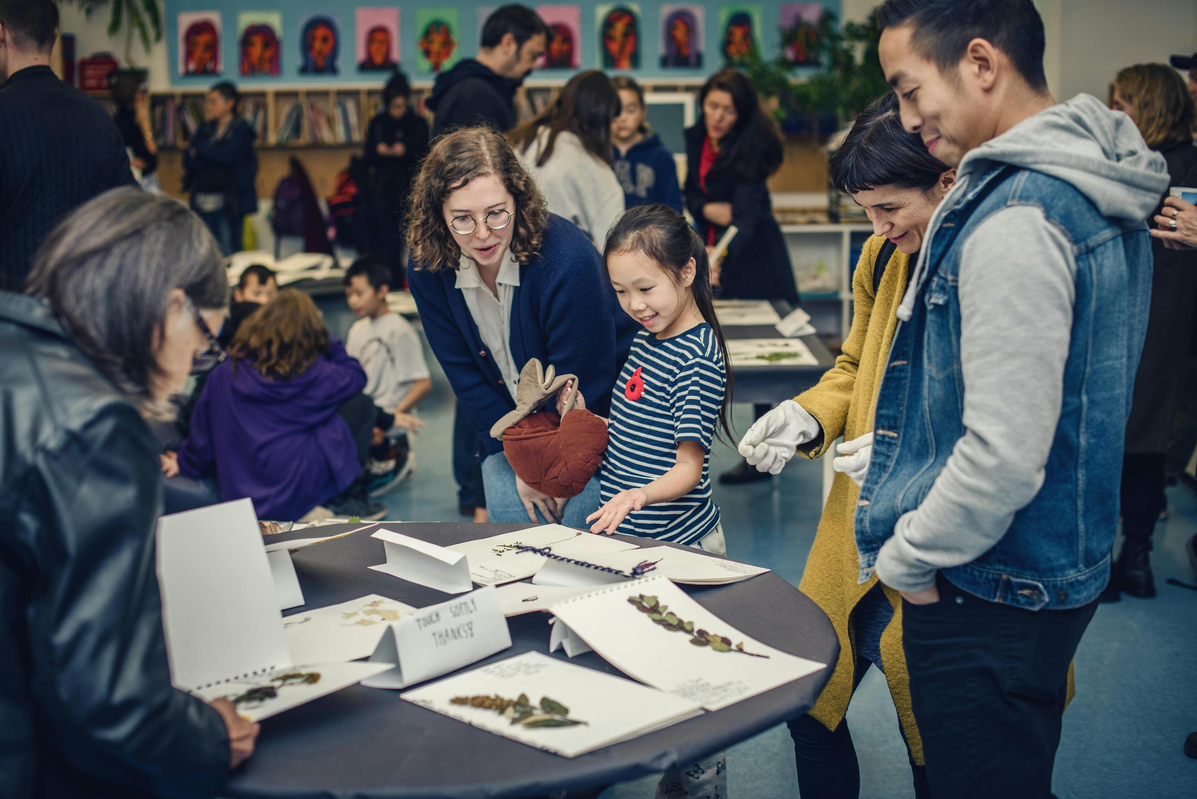 In a classroom, children and adults gather around black round tables to see students’ artworks placed on them. The works are paper-based, on which plants and some writings are attached. 