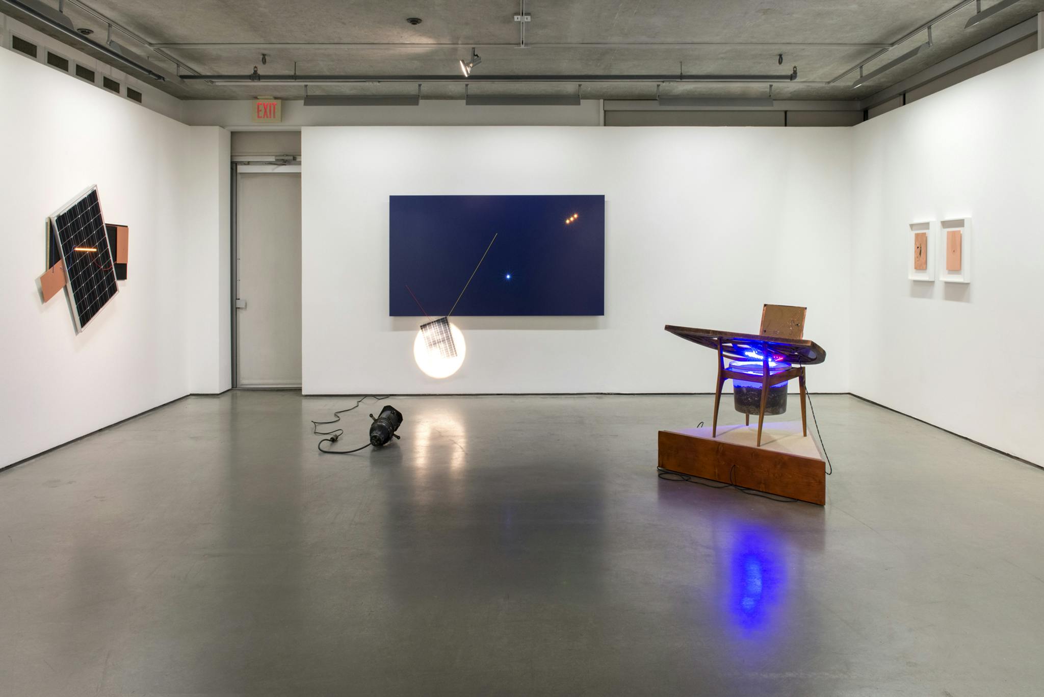 Various multimedia works are installed throughout a gallery. A sculpture resembling a desk chair on a triangular plinth foregrounds three multimedia artworks hanging on three gallery walls.