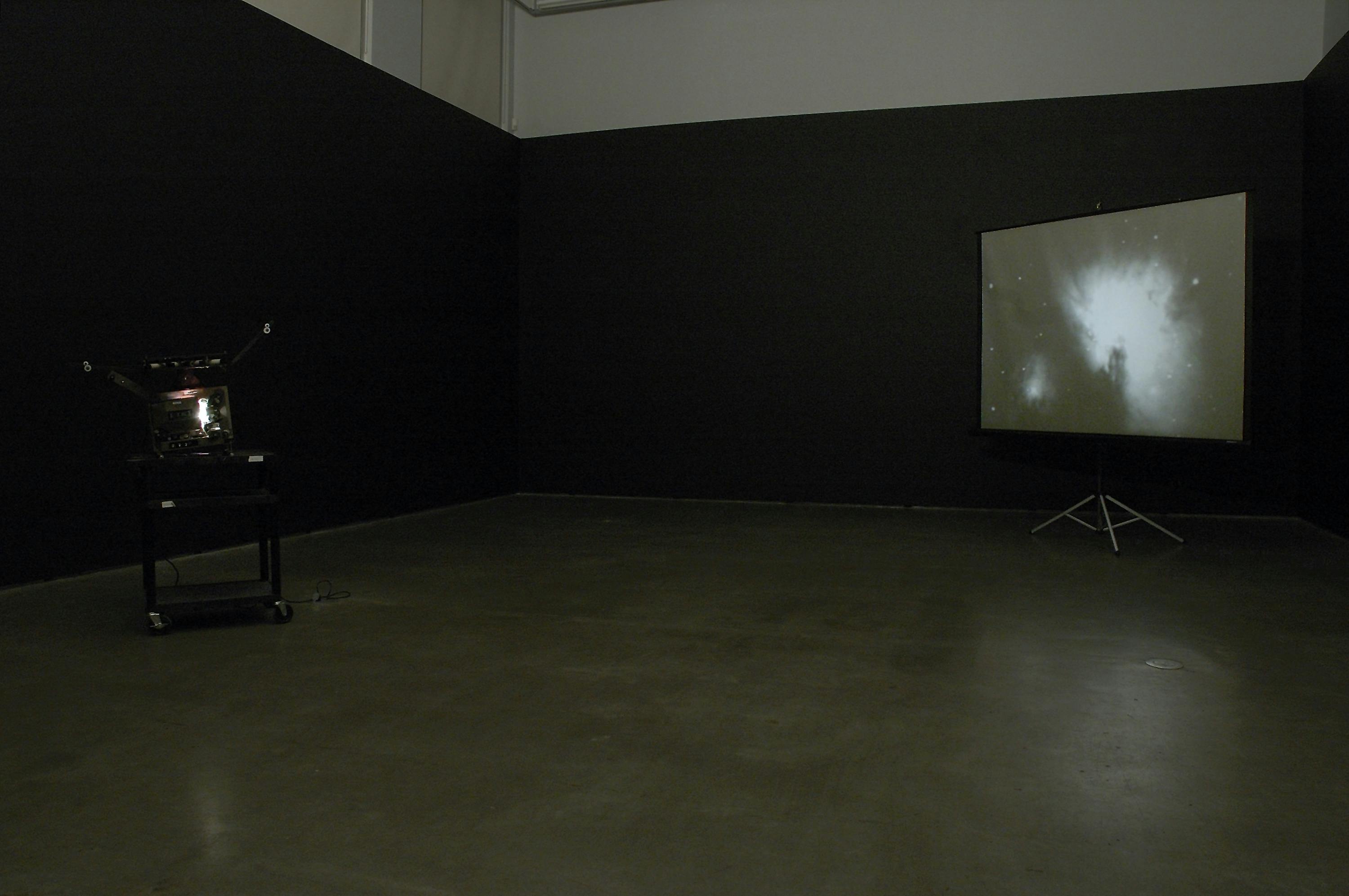 A film projector is installed in a darkened gallery space. A projector placed in a corner of the room shows a moving image of outer space. White-blue stars and a nebula are depicted in the image. 