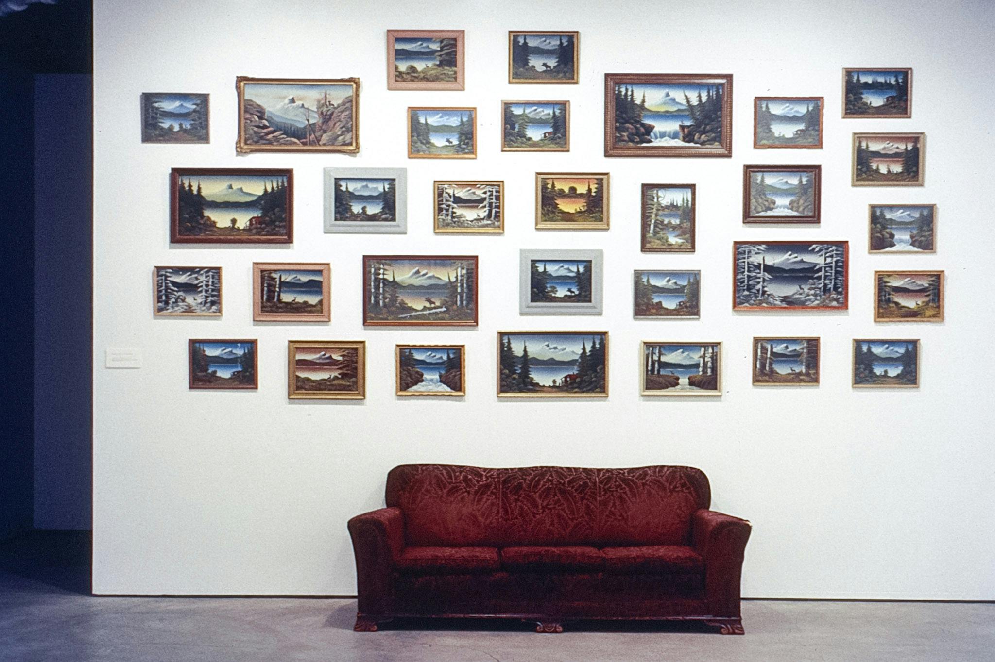 31 landscape paintings of many sizes in various frames mounted on a white wall in a gallery space. Below, in front of the wall, there is a faded red velvet couch with a feather-shaped paisley pattern.