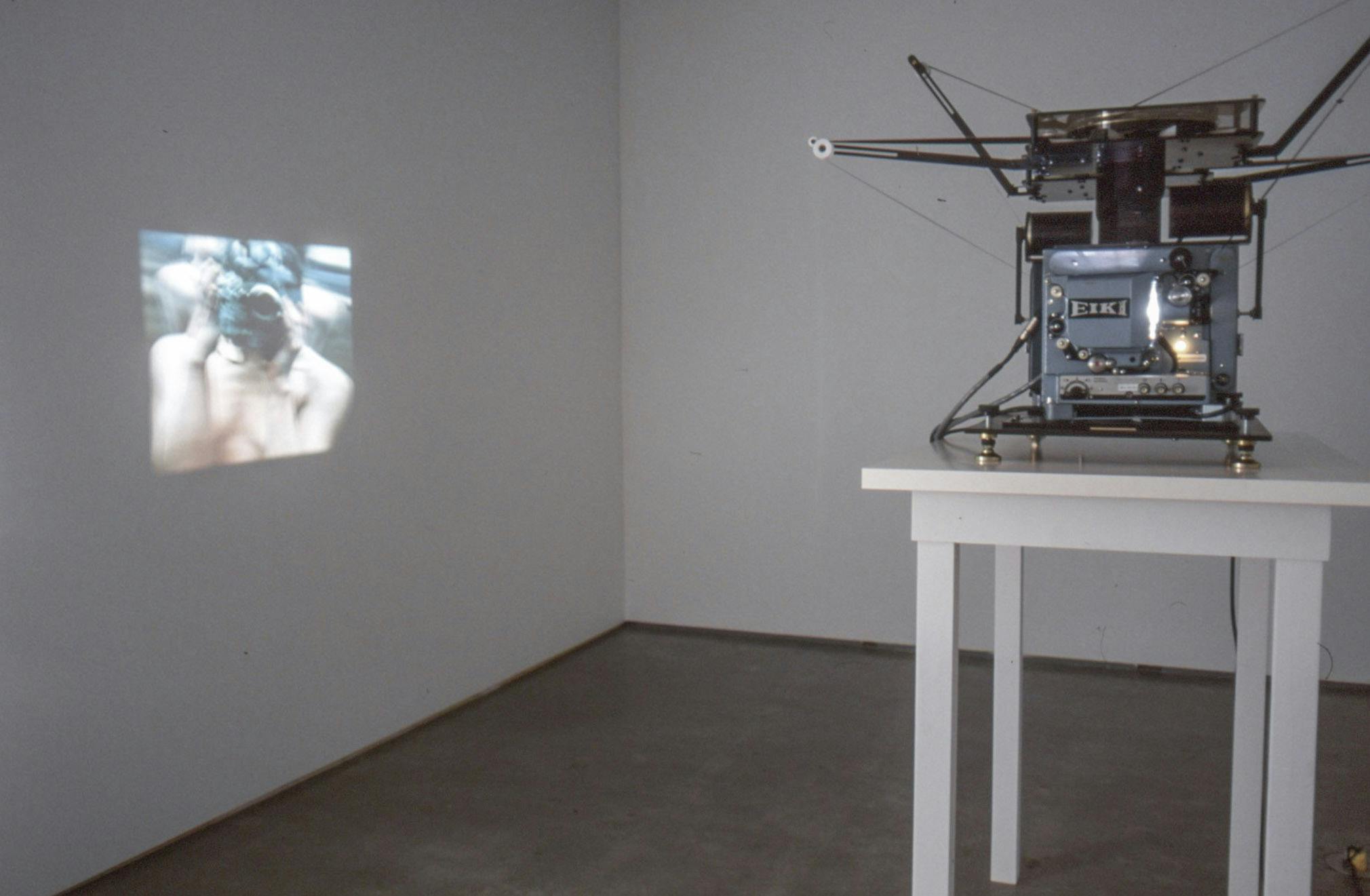 A silver film projector is placed on a white table. It projects a film on a gallery wall. The projected blurred image depicts a hand and a green coloured large insect-like shaped object. 