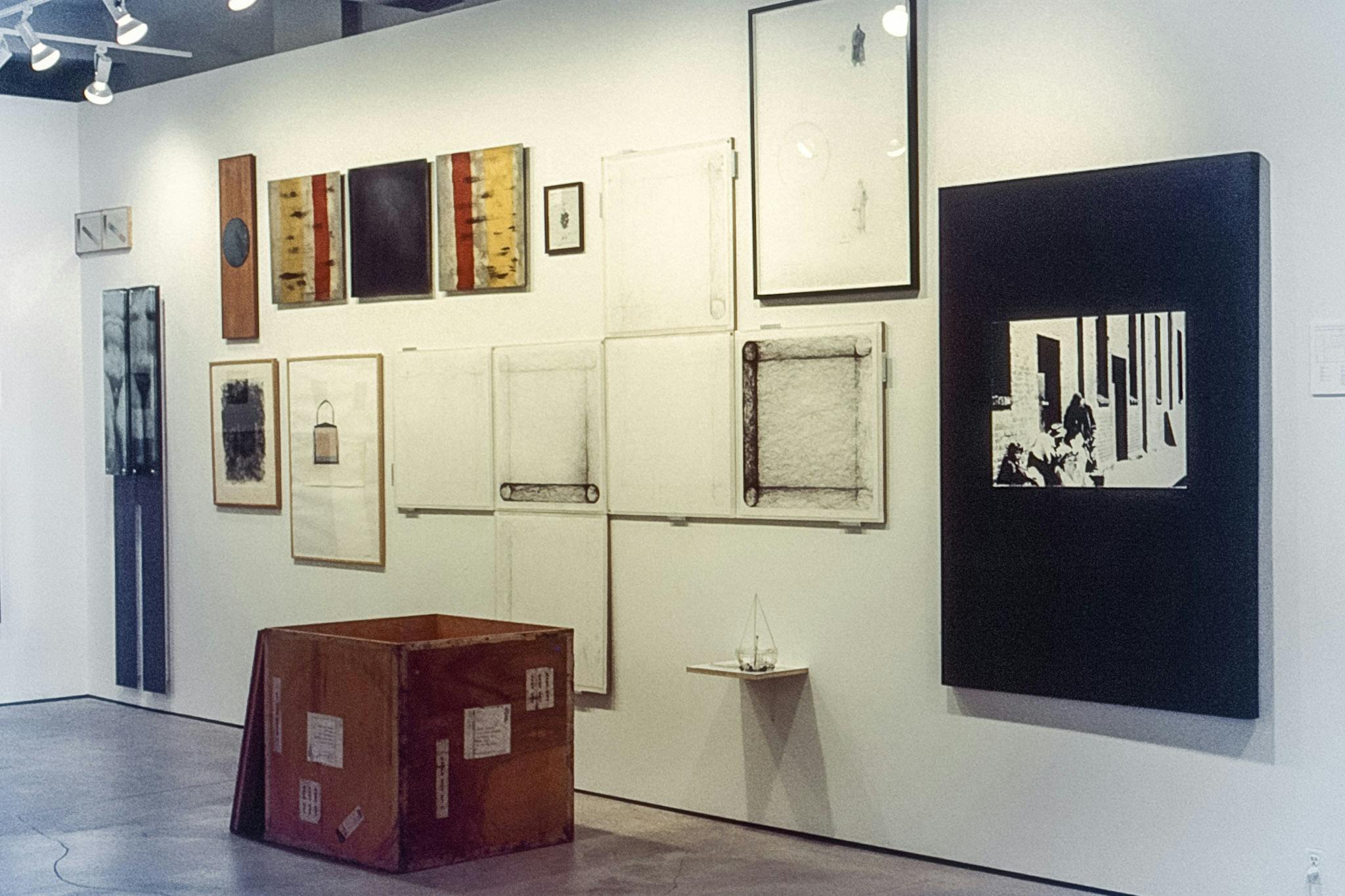 A photo of various artworks mounted on the gallery wall. Most of the paintings and photographs are black and white. There is also a large wooden box with multiple labels sitting in front of the wall. 
