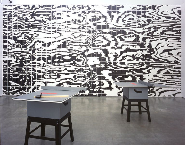 Two small grey desks are placed in a gallery space. The gallery wall behind the tables is covered with a black and white abstract pattern that looks like an enlarged photograph of a TV screen. 