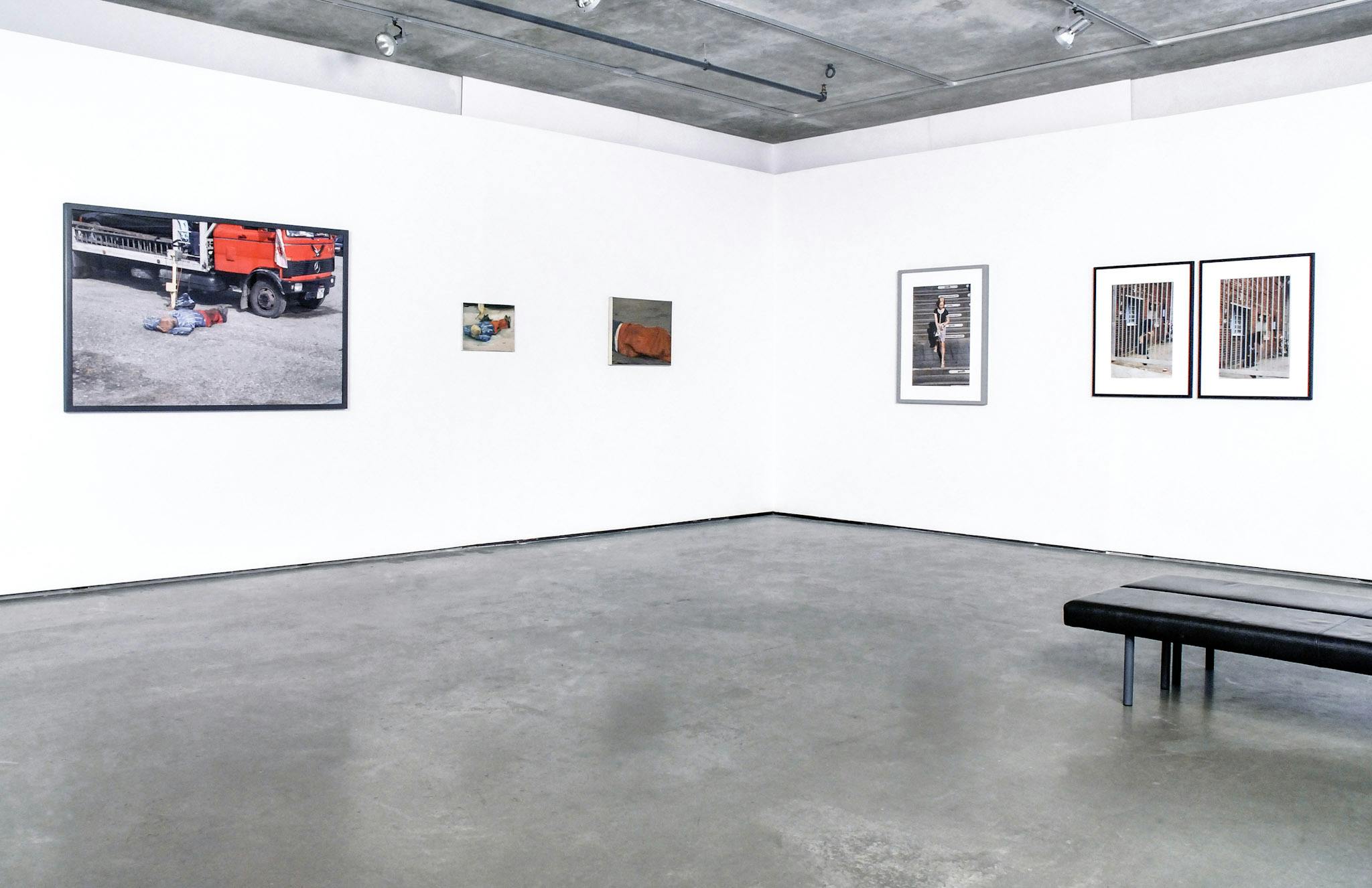 Six photographs are installed on the gallery walls. Two of them are framed photographs depicting a shutter gate. A large photograph on the left depicts a mannequin beside a fire truck. 