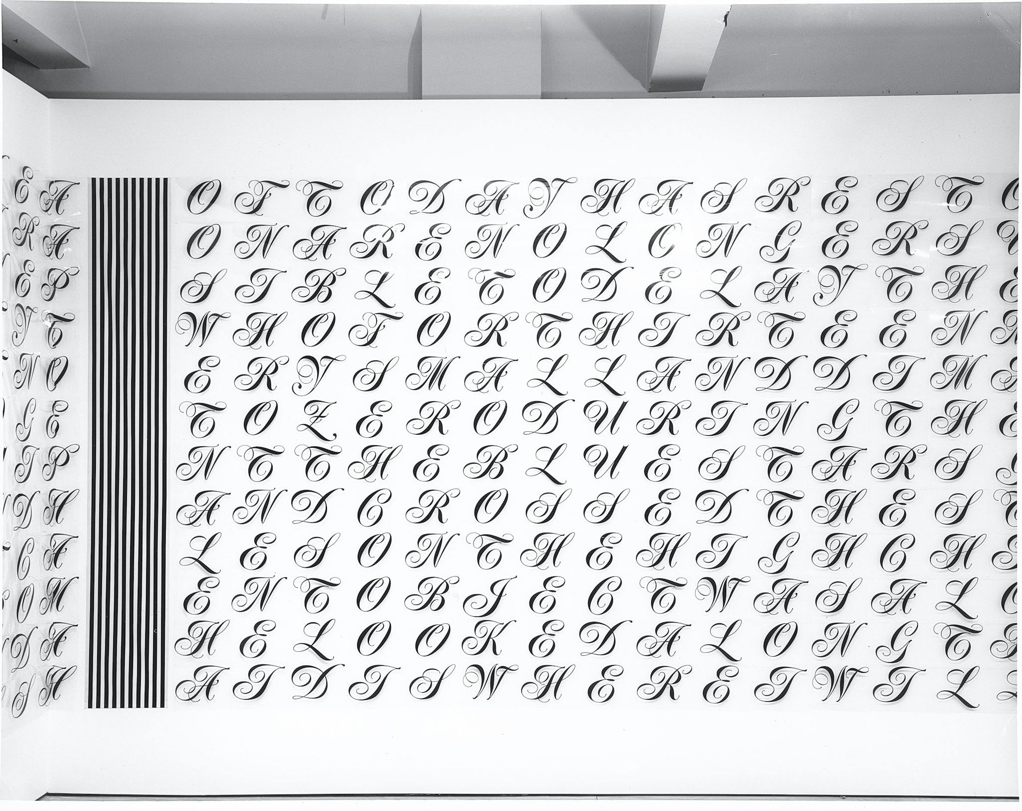Cursive capital letters mounted on a white wall. They are printed on clear plastic sheets and are arranged neatly but out of alphabetical order. The letters are thin and highly ornate. 