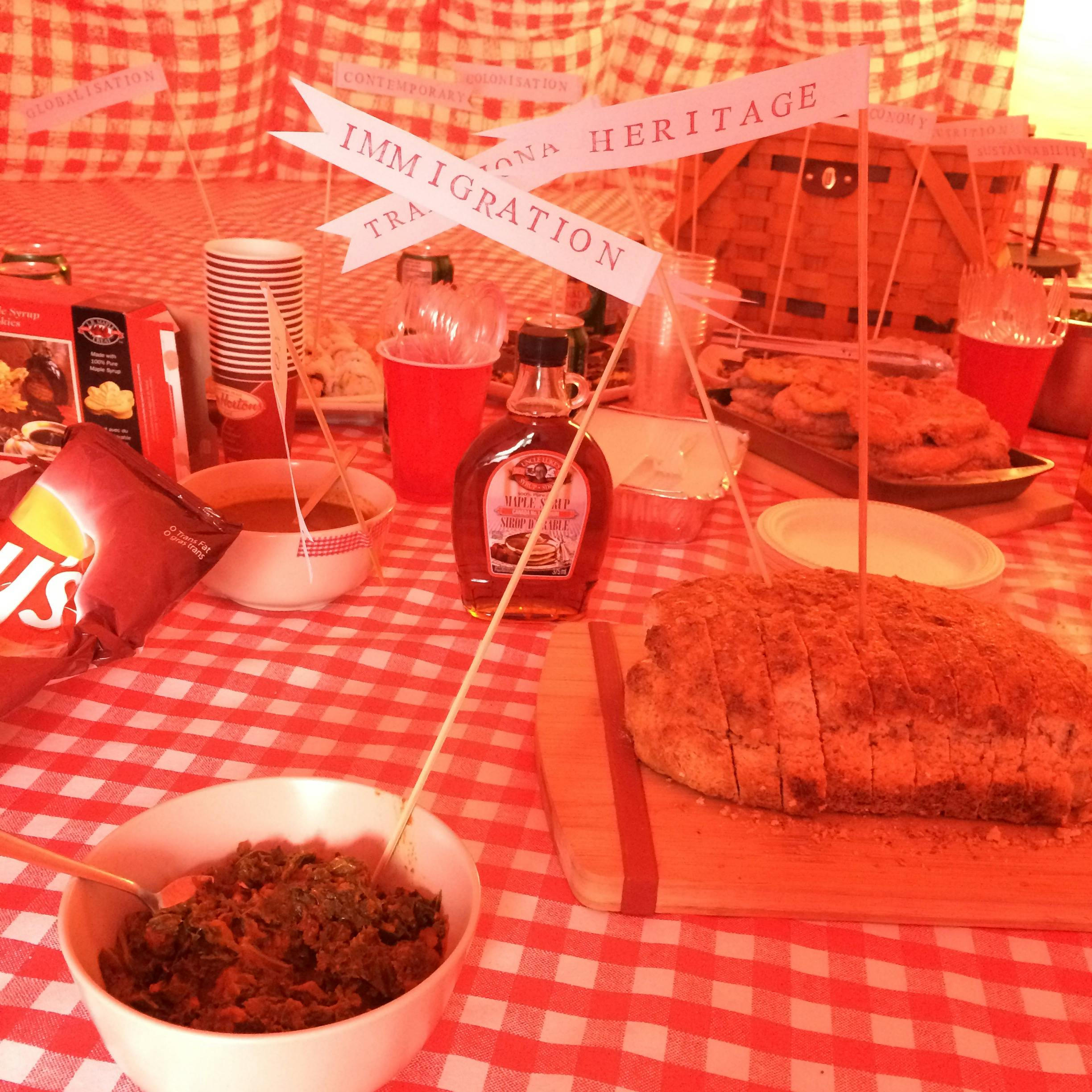 An image of many dishes placed inside red and white gingham patterned tent. Small flags showing various words, such as immigration and heritage, are sticking out of each dish.