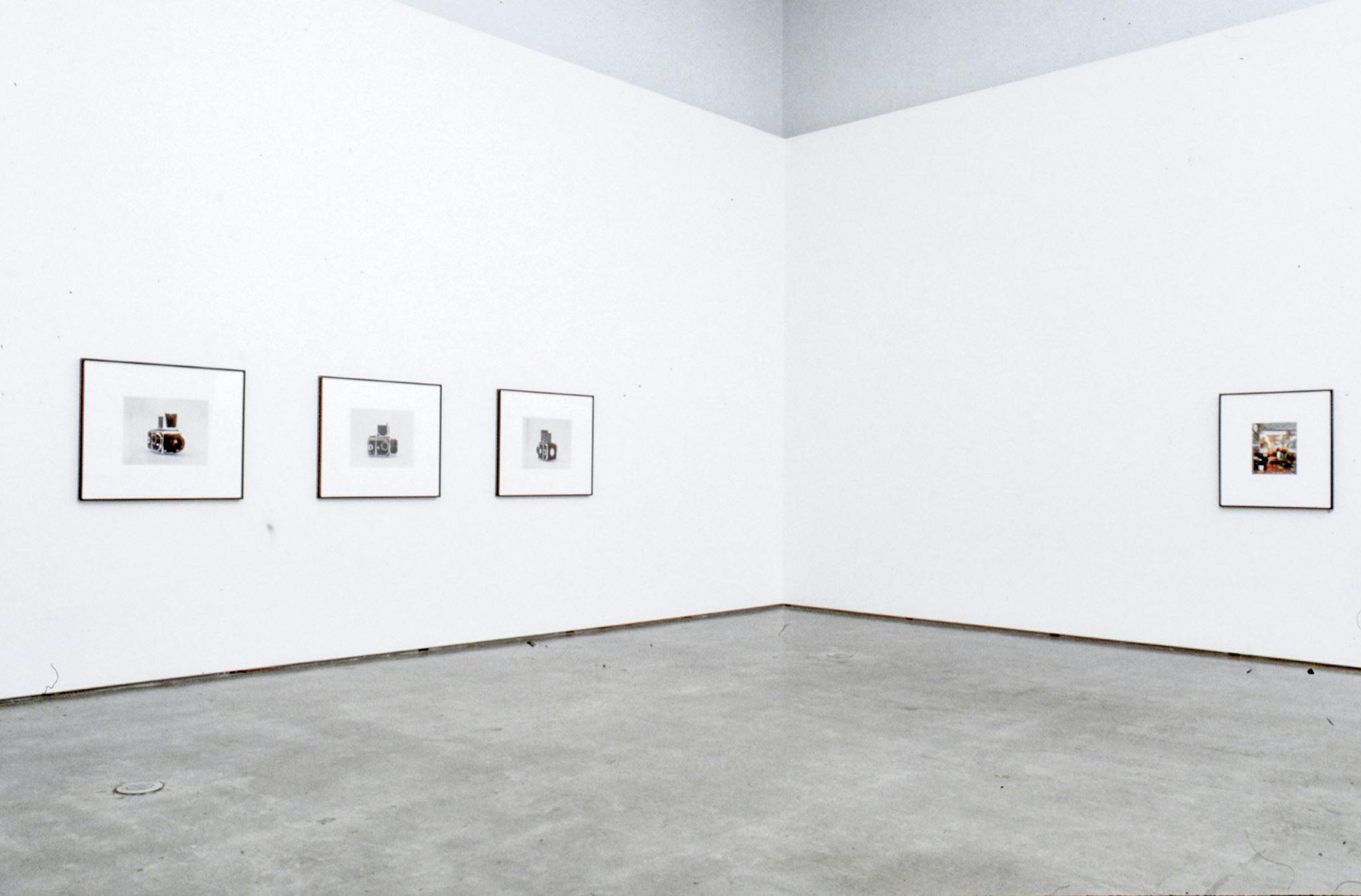 An installation image of coloured photographs on gallery walls. Three photographs of black old cameras are on the left wall. The photograph on the right-side wall shows some round orange objects.
