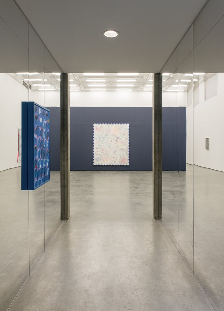 A hallway covered in mirrored panels leads into a bright gallery. A blue, abstract painting hangs on one hallway wall. A large-scale abstract painting is visible on a blue wall inside the gallery. 