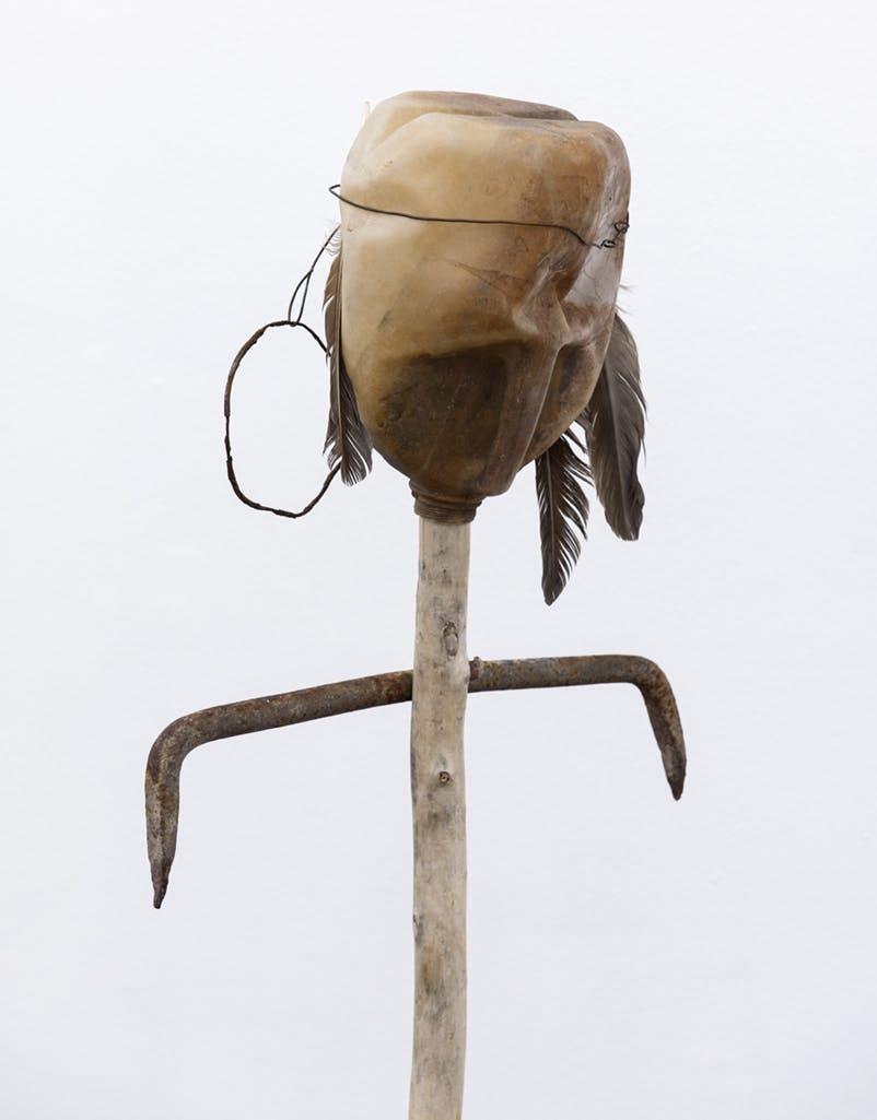 A sculpture made from a plastic milk jug stuck upside down on a wood stick, from which arm-like metal rods extend out horizontally. Feathers attached to a wire band hang from the upper part of the jug.