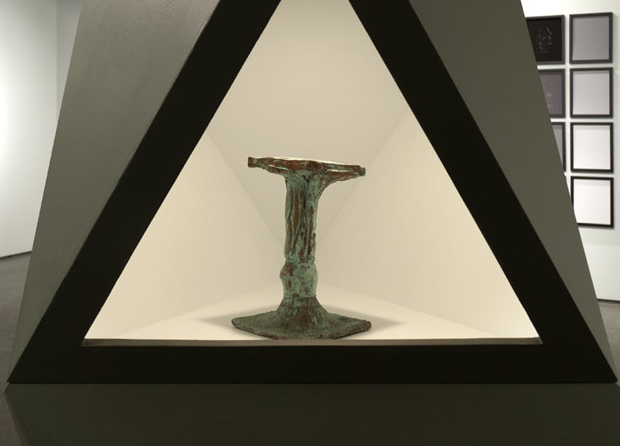 Detail image of a geometric sculpture. Within the sculpture, a lit, hollowed out triangular space houses a relic-like object.