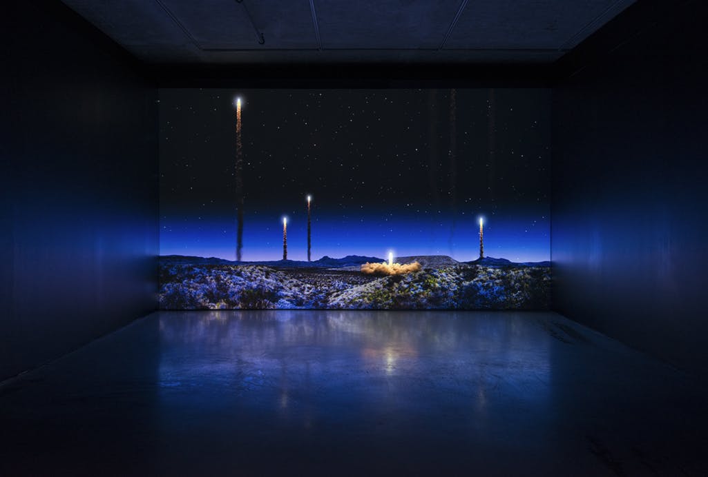 A single-channel video is projected on a gallery wall. The video depicts objects launching from a desert landscape into a starry night sky, only their path of light and smoke is visible.