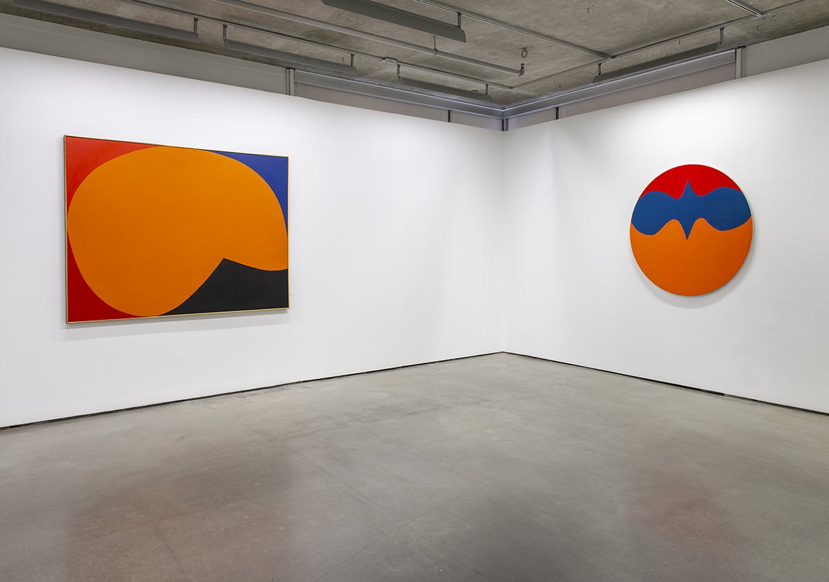 Two paintings by Leon Polk Smith sit on either side of a corner in a gallery space. The paintings are made up of simple, bold shapes in colours such as orange, red, blue and black.
