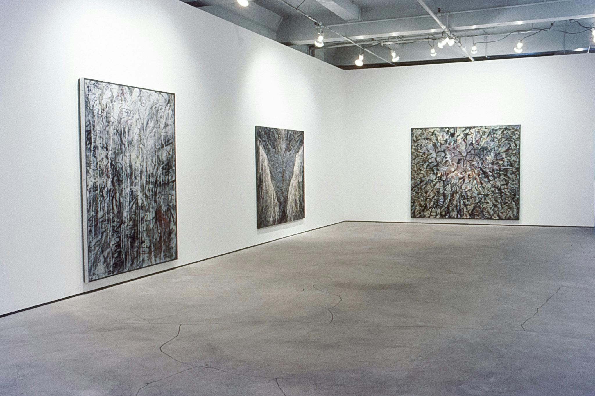 3 paintings on large canvases of different proportions, in a gallery. They have a splattered and swirled paint with different materials. The painting in the centre has an inverted triangle form in it.