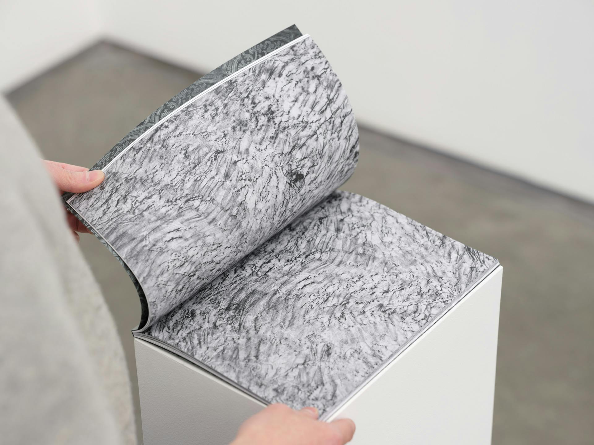 A thin artist book on a white plinth. A pair of hands hold a page open to show facing pages with abstract grey texture printed allover.