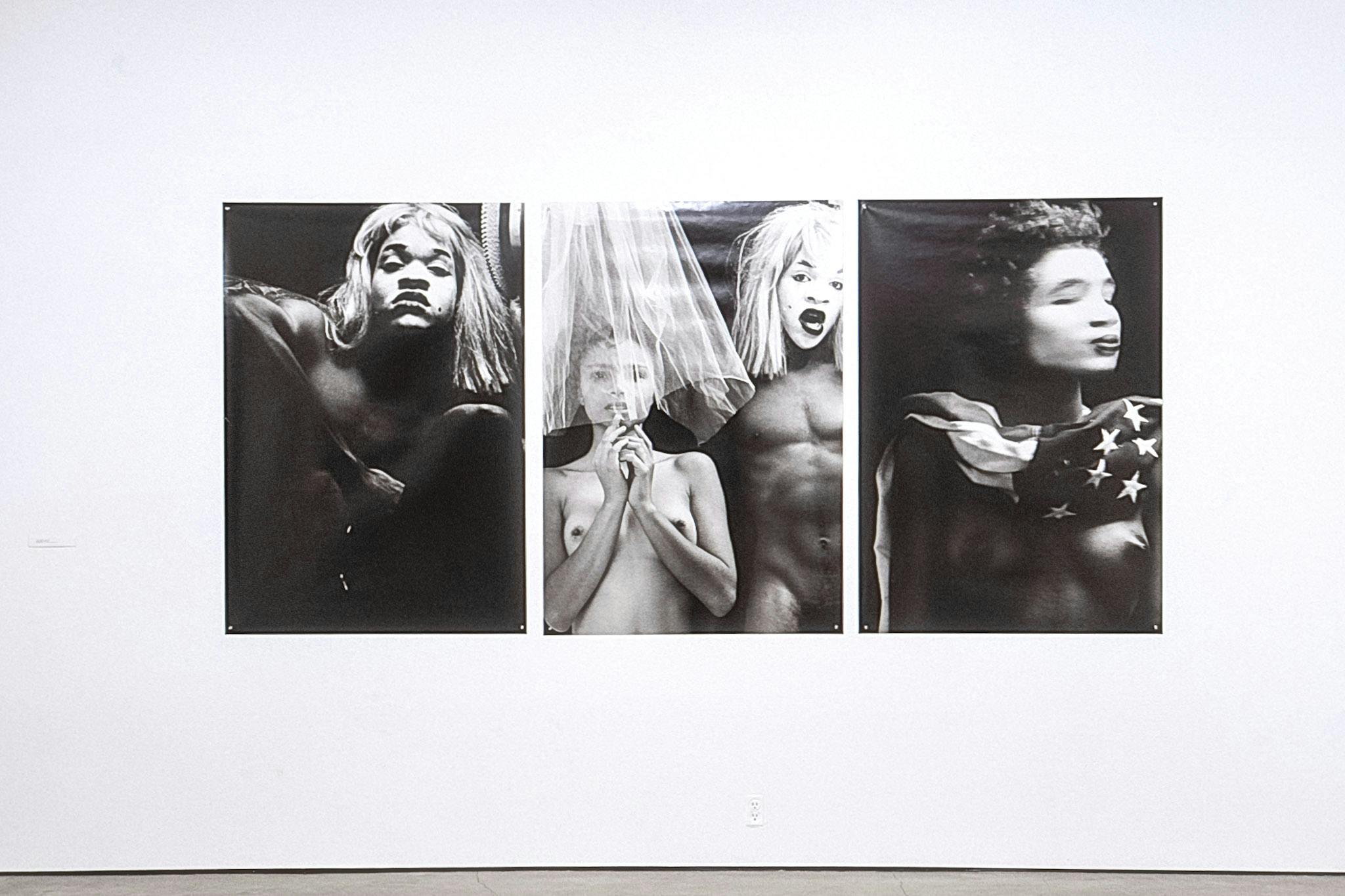 Three large photos on a gallery wall. The photos are black and white and show semi-nude people in drag or costume. The photos are printed on glossy paper and pinned to the wall.