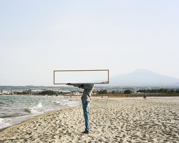A photograph of a person standing on a beach near the ocean. The person is holding a rectangular mirror horizontally with one arm outstretched. The reflection in the mirror is of a light blue sky.
