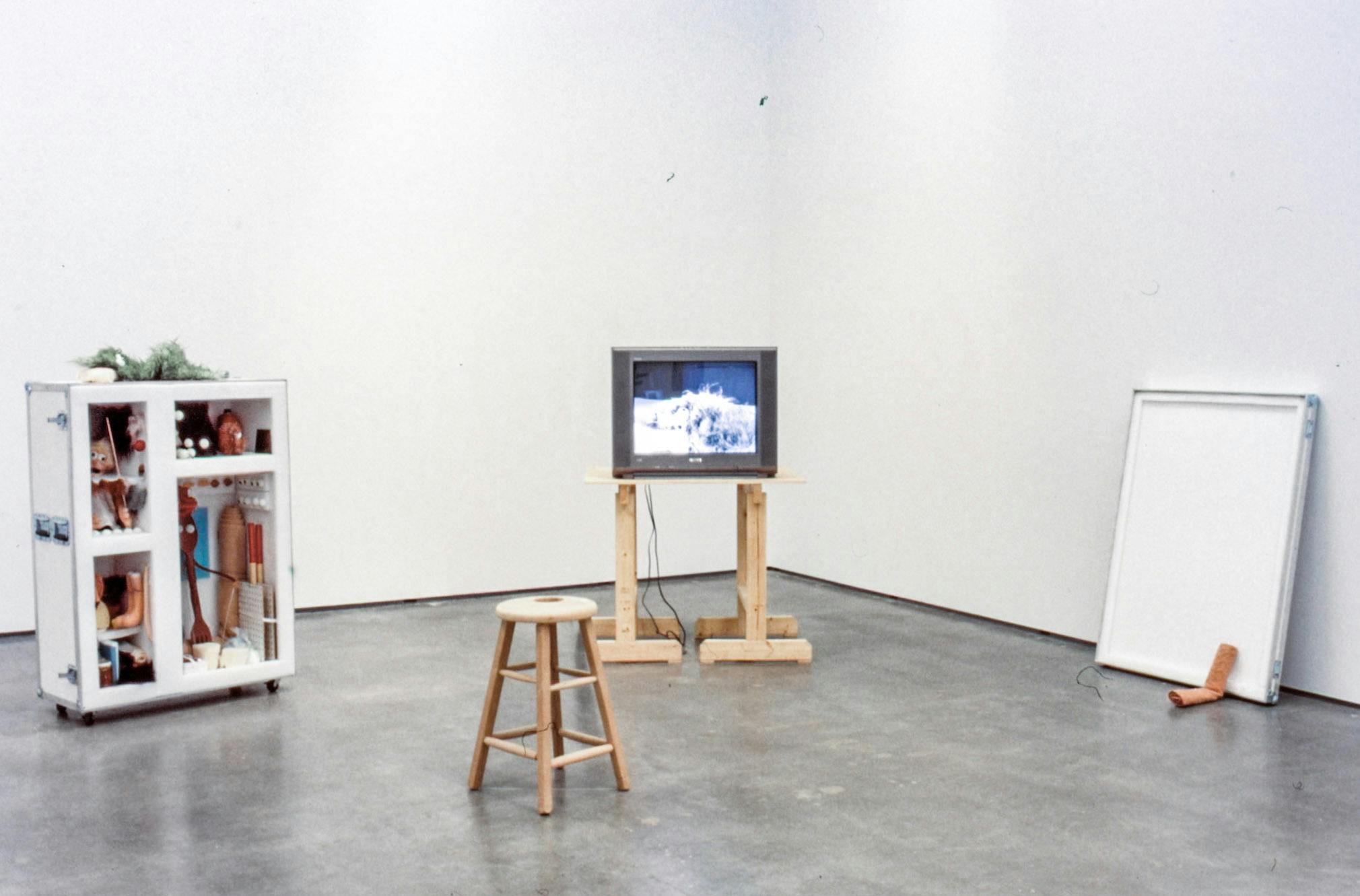 In a corner of a gallery room, a CRT TV is placed on a wood coffee table. A video work is shown on that TV. A mobile storage cabinet filled with objects is placed next to the TV. 