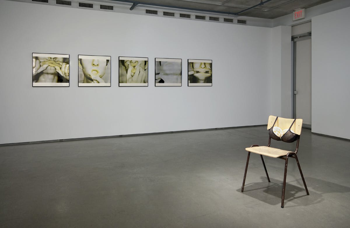Five close up images of a mouth and hands hang on the wall of a gallery. A chair sits off to the side with a bra attached to the back support. The bra is holding two round objects. 