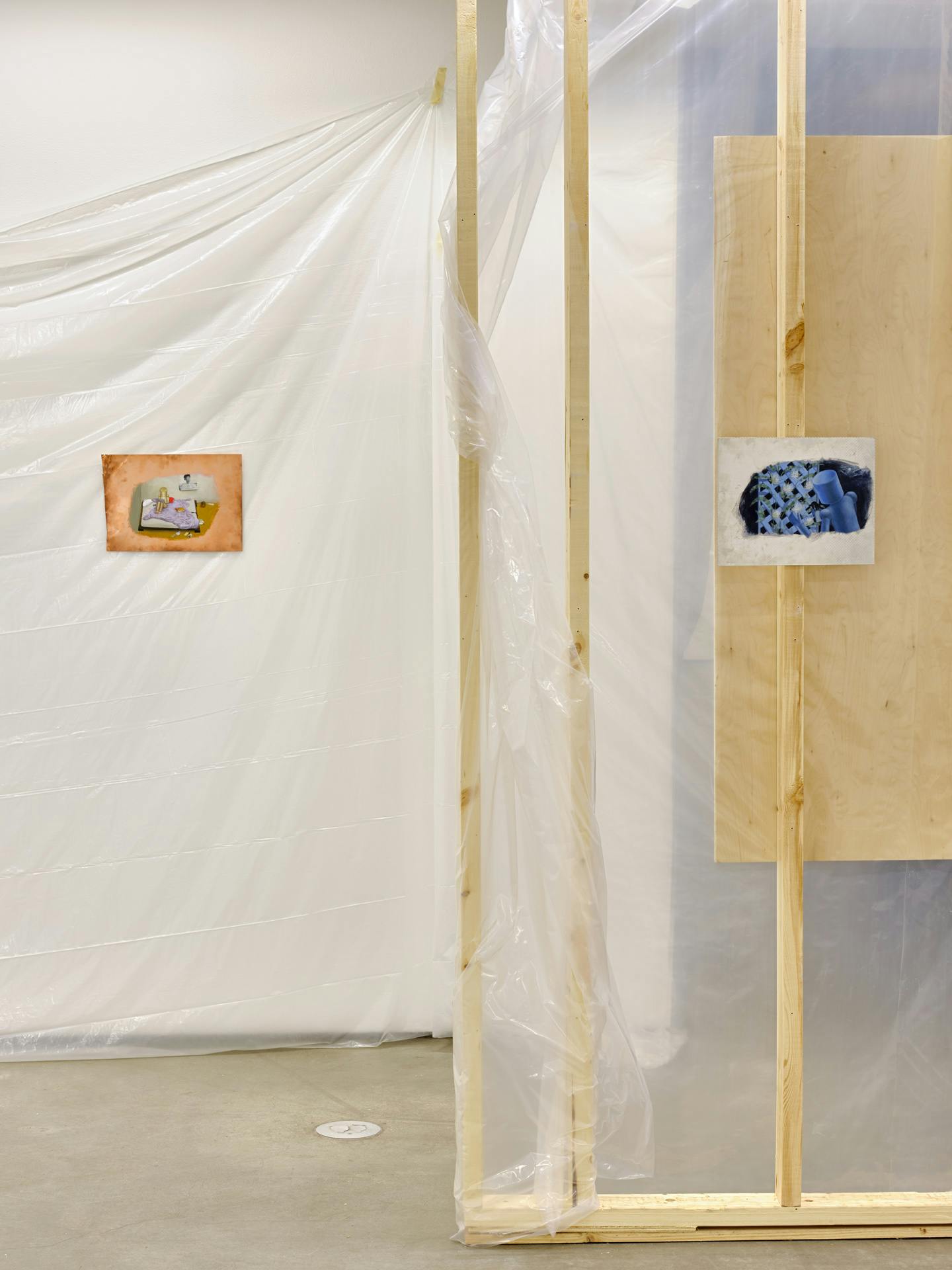 A small copper painting hangs on a white gallery wall draped in translucent plastic sheets. To the left a small blue painting on a metal sheer hangs on an unfinished wooden wall structure.