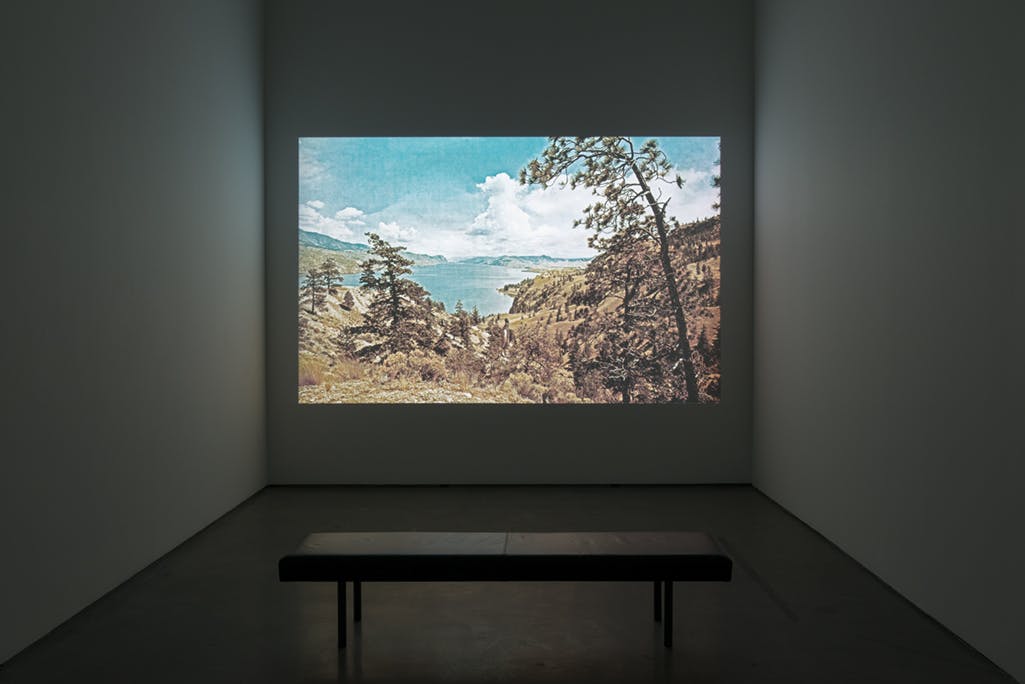 A single-channel video is projected on a gallery wall. The video depicts a hilly, rocky landscape dotted with evergreen trees surrounding a blue lake. 