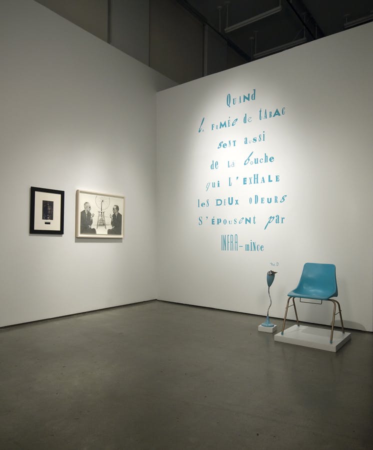 Two framed photographs hang side by side on the left side of a corner wall. On the right side, large blue french text is written on the wall. By the same wall, a blue chair and sculptural object sits.