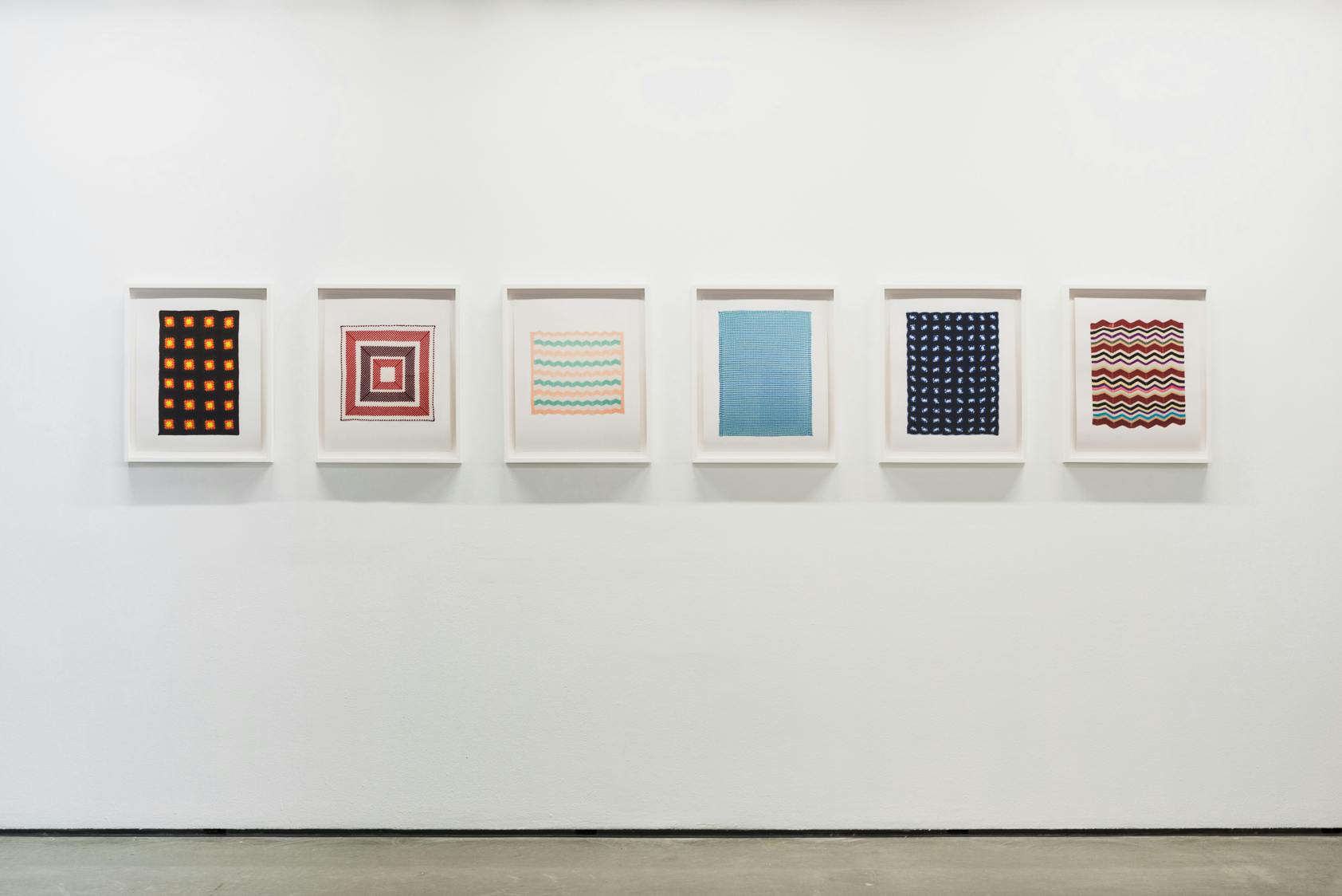 Six works from Brent Wadden’s exhibition Two Scores hang in white frames on the wall of a gallery space. They are photographs of woven afghan blankets of different patterns and colours.