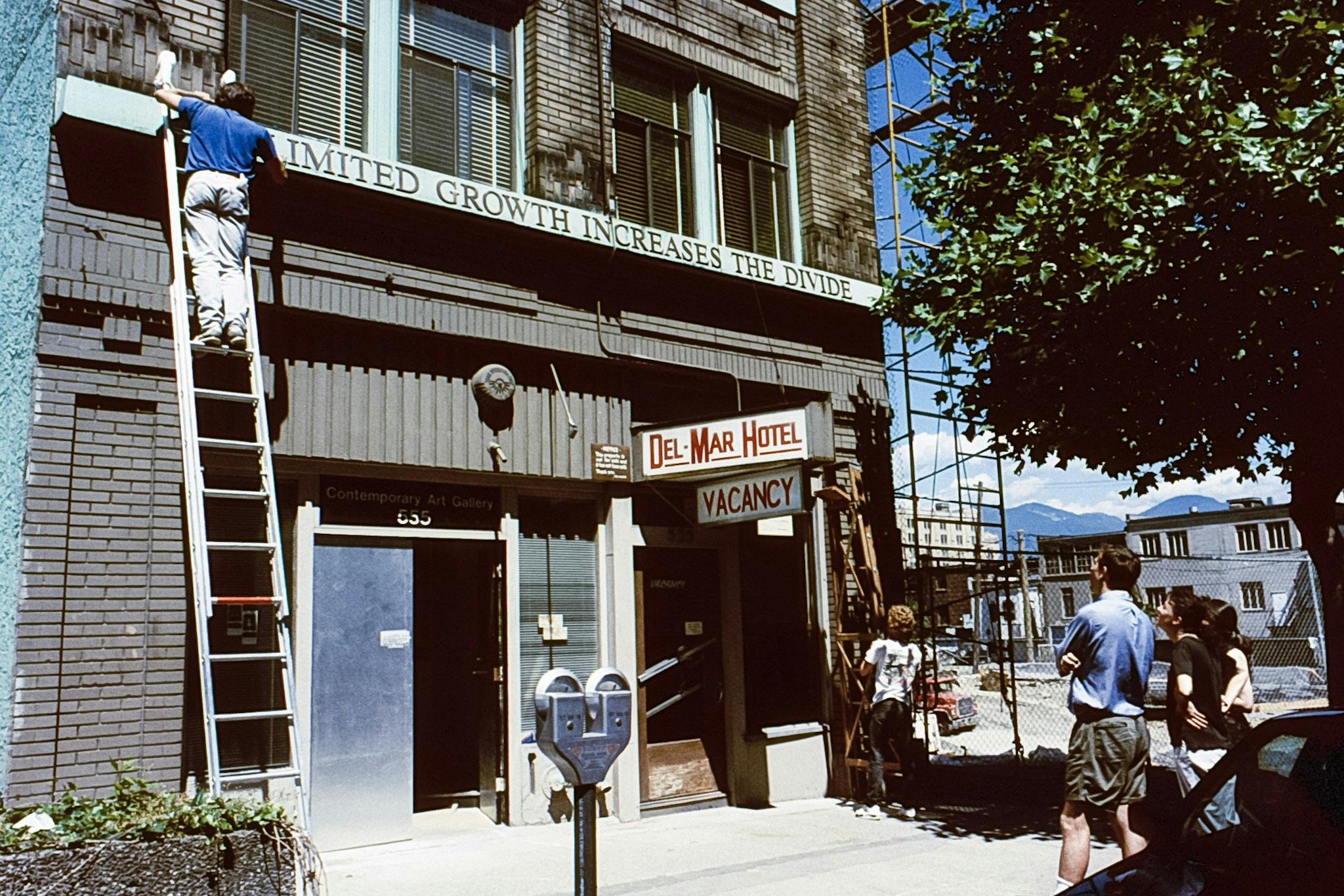 On the front of a building, 2 people working on ladders install metal letters. They read: "Unlimited Growth Increases The Divide." While the two people work, three others stand outside, looking on. 