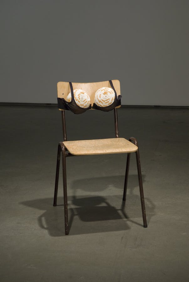 A wooden chair is installed on the grey floor. A black bra is clasped on the back support of this chair and holding two large balls, which are covered by rolled white and orange paper. 