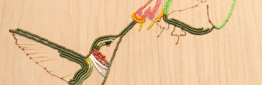 An image detail of a wood veneer panel with a rendering of a hummingbird drinking nectar out of a flower. The rendering is composed of bead-embroidered and wood-burned lines.