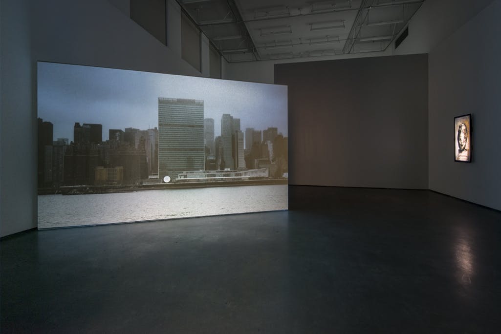 A single-channel video is projected on a large screen in a gallery. The screen is installed at an angle, jutting out to the right from the wall. On screen, a city skyline rises above a body of water.