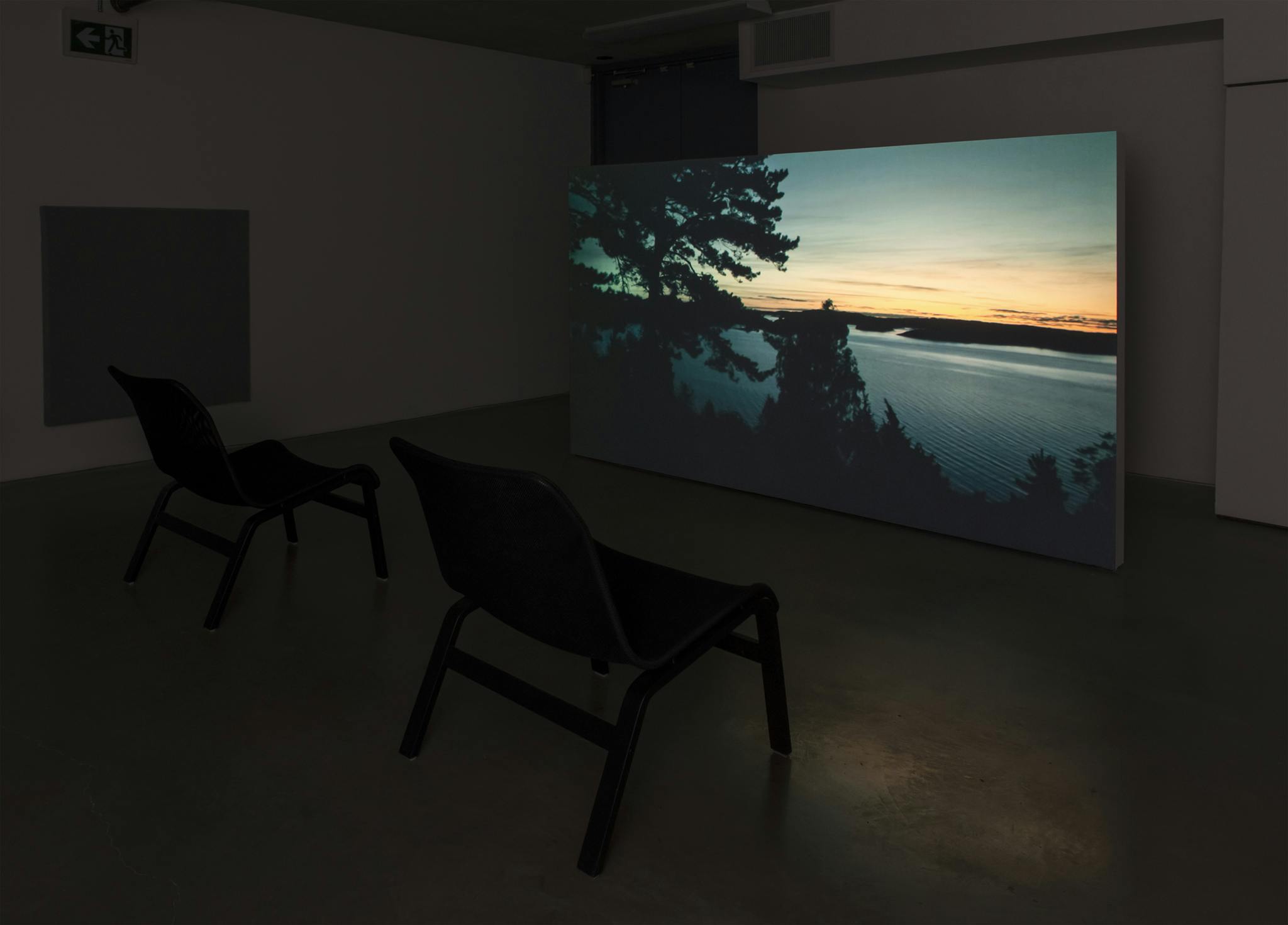 A dark room with two chairs facing a large video projection. The projection is a still image of a sunset through the trees, looking over the water. 