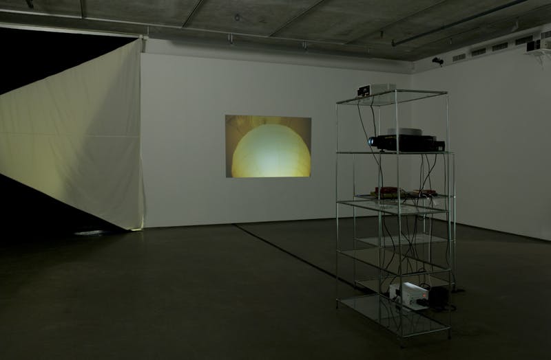 In a darkened gallery space, two glass shelves sit in the middle of a gallery, holding slide projectors at various heights. An image of a white, round-shaped light is projected on the wall.