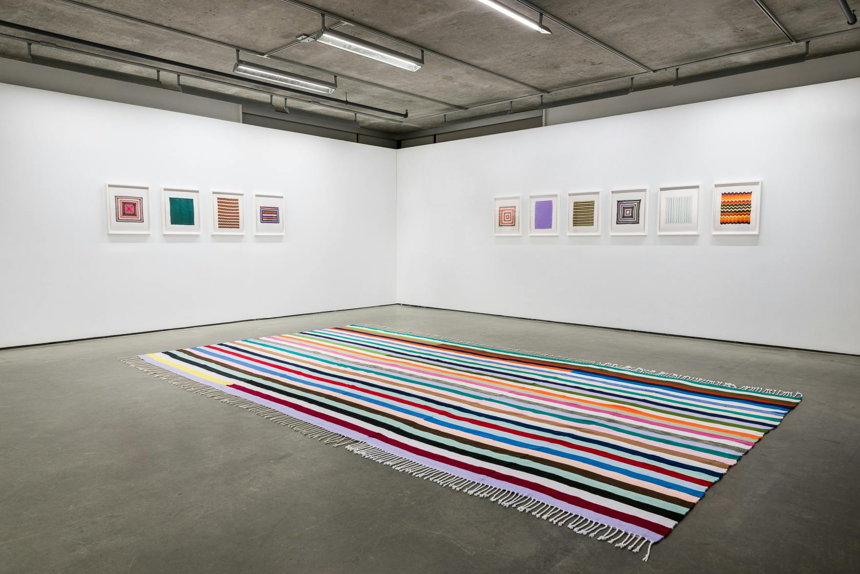 Installation image of Brent Wadden’s exhibition Two Scores. Four framed photographs hang on the left side of a corner wall and six on the right. A large scale woven rug lays on the floor. 