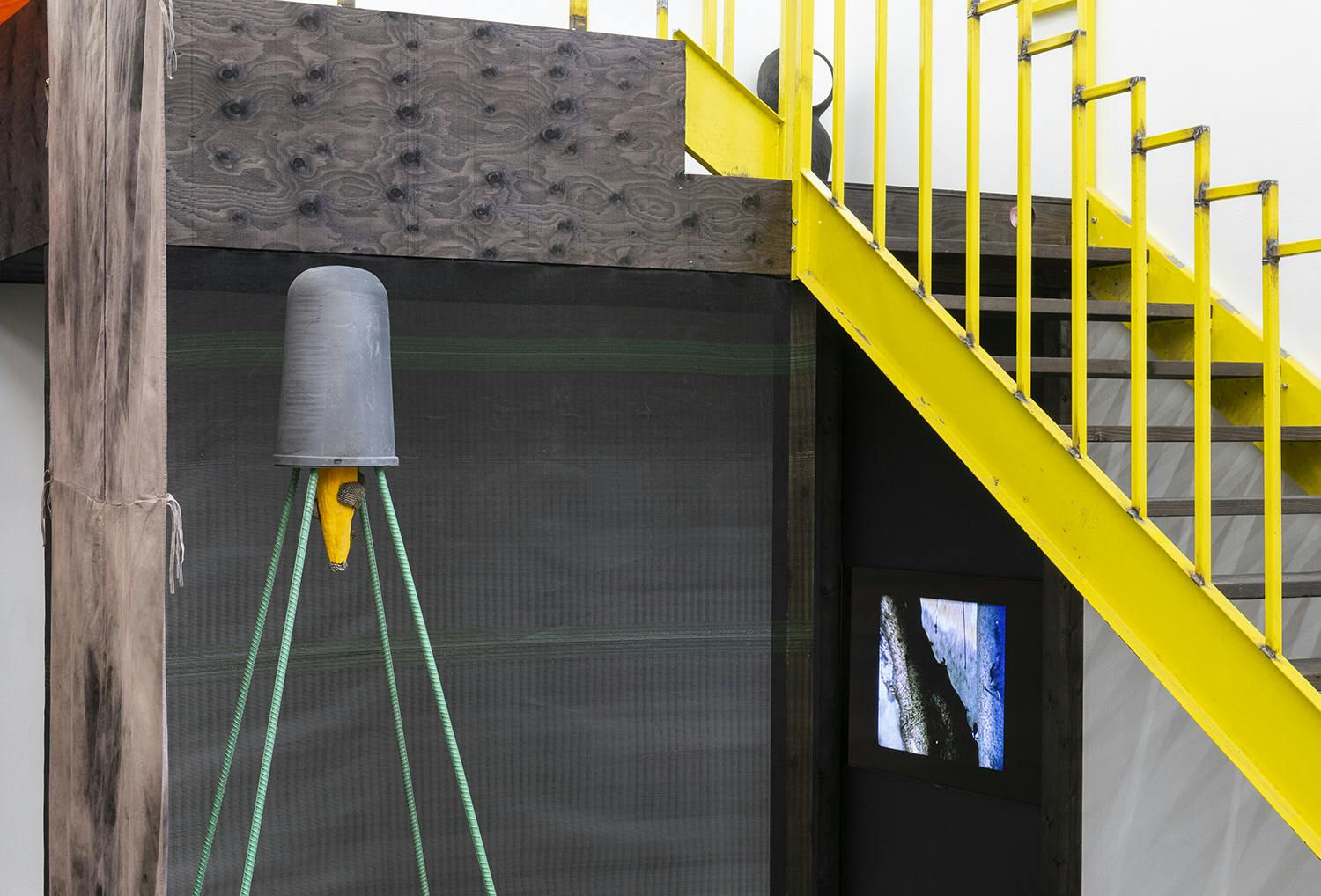 Yellow stairs go up to a platform on the right side of the image. Under the stairs there is a monitor showing an abstract picture. On the left side of the image there is a tall sculpture. 