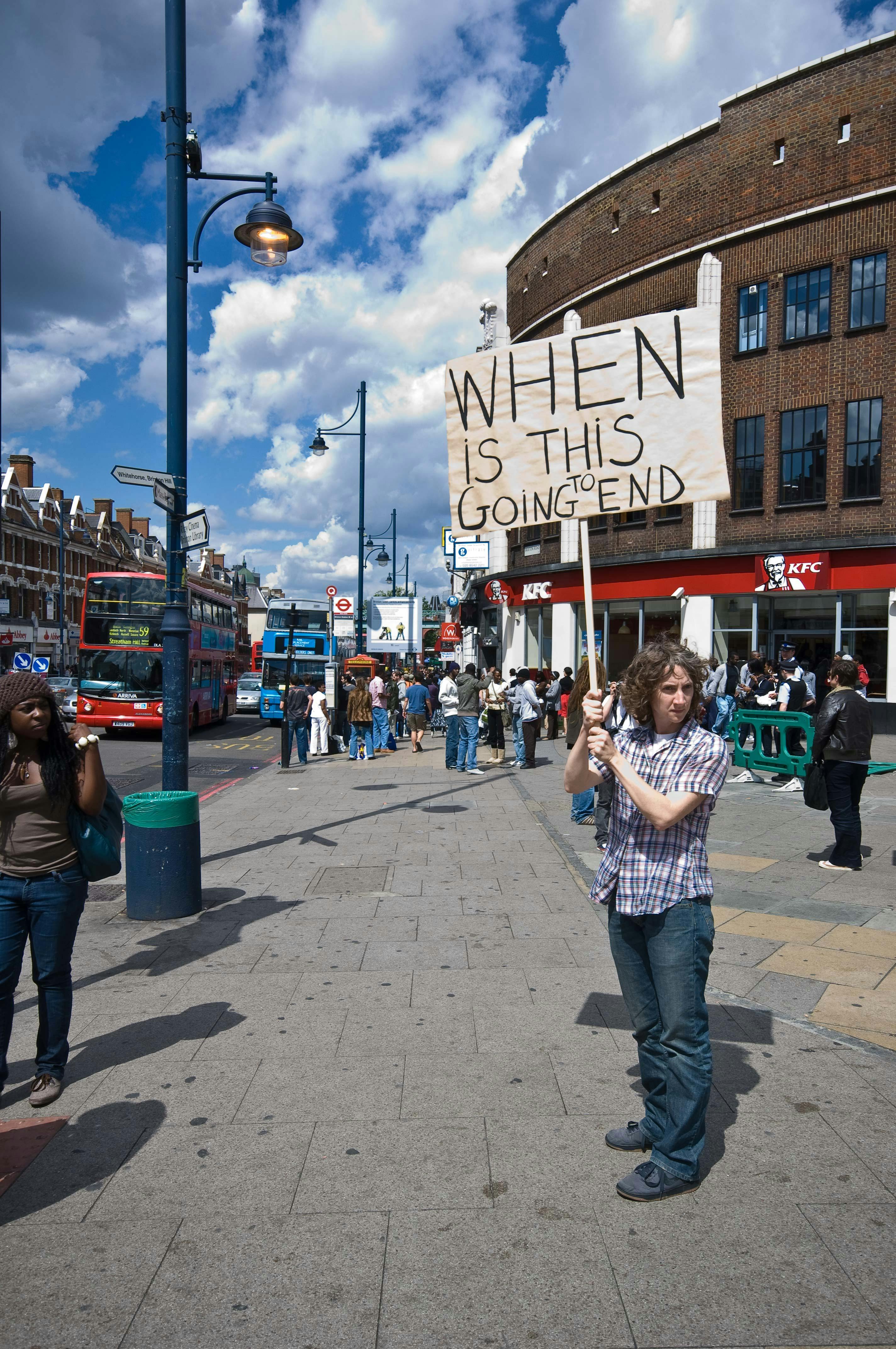 A photograph depicting a protester on a city street holding a sign that reads "WHEN IS THIS GOING TO END."  