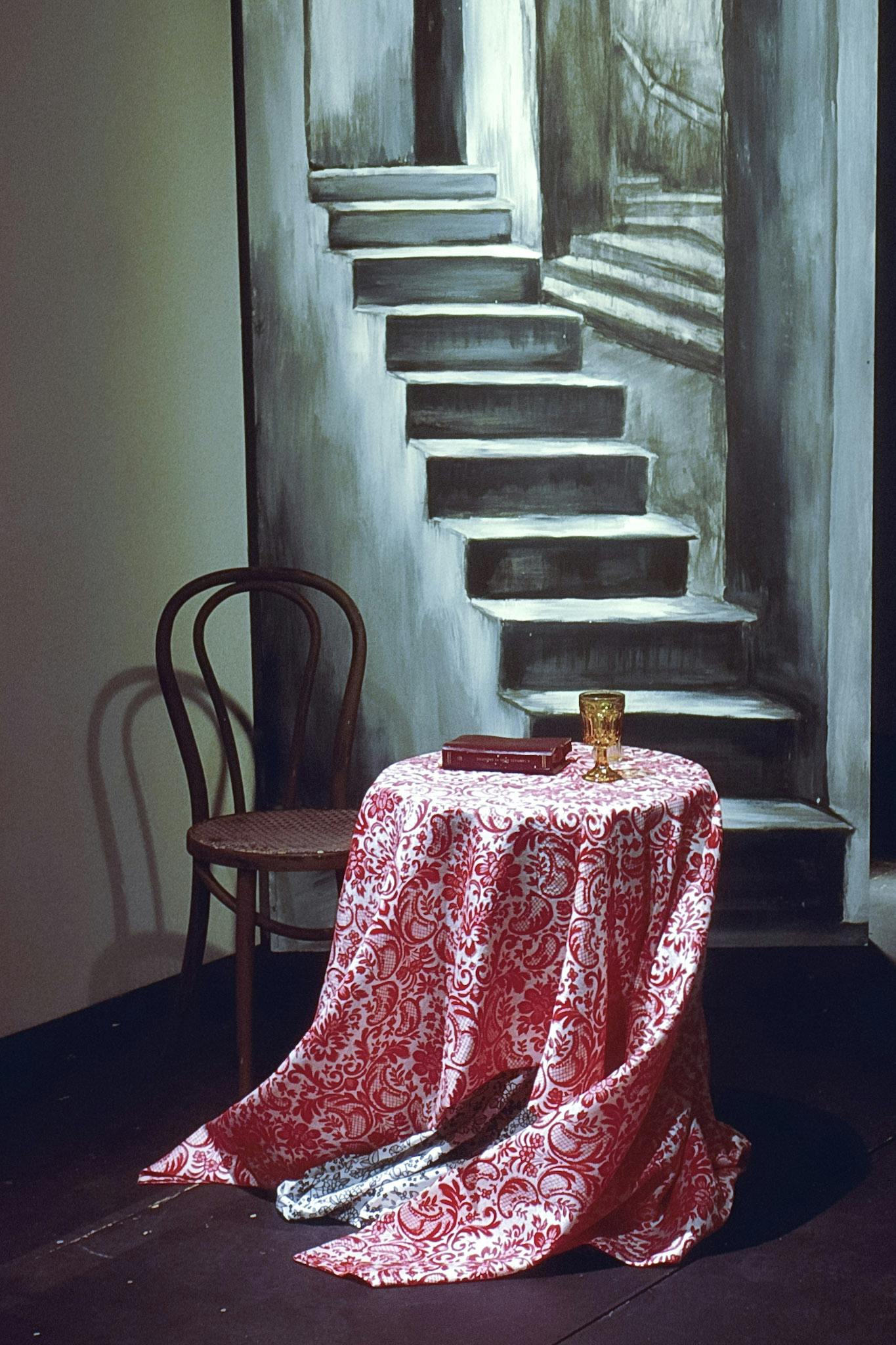 A small round table and a wooden chair are on a corner of the theatre stage. A red botanical-patterned tablecloth covers the table, on which a vintage wine glass and a book are placed.